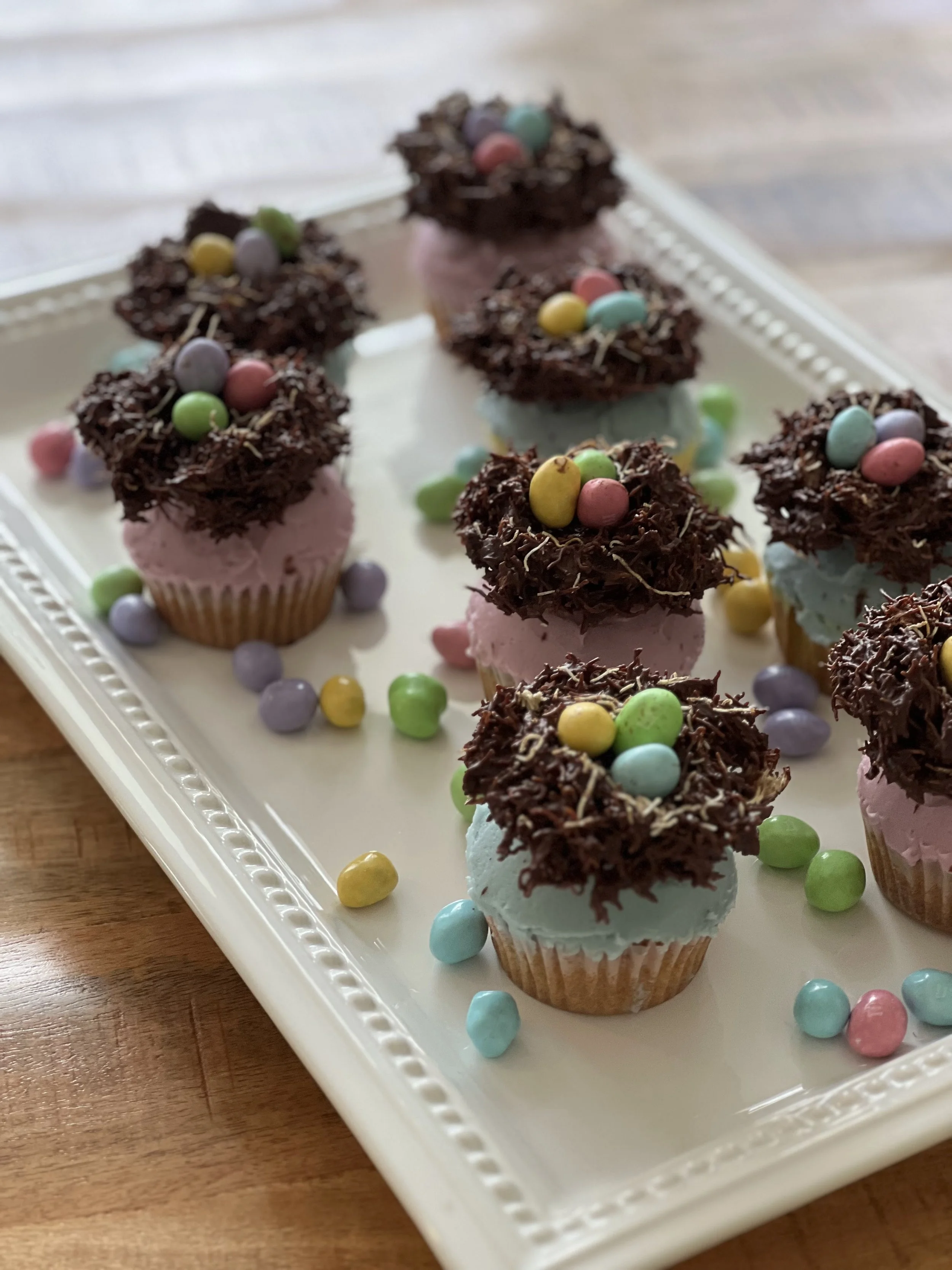 Decorated cupcakes with pink and blue frosting, topped with chocolate shavings and colorful candy eggs, arranged on a white tray.