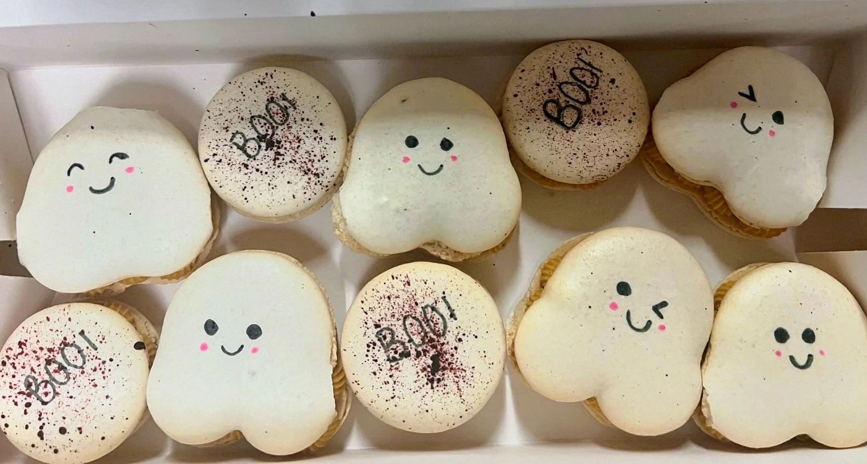 A box of decorated cupcakes with smiling face, winking face, and "Boo!" designs, some with cocoa powder and eye and mouth drawings.