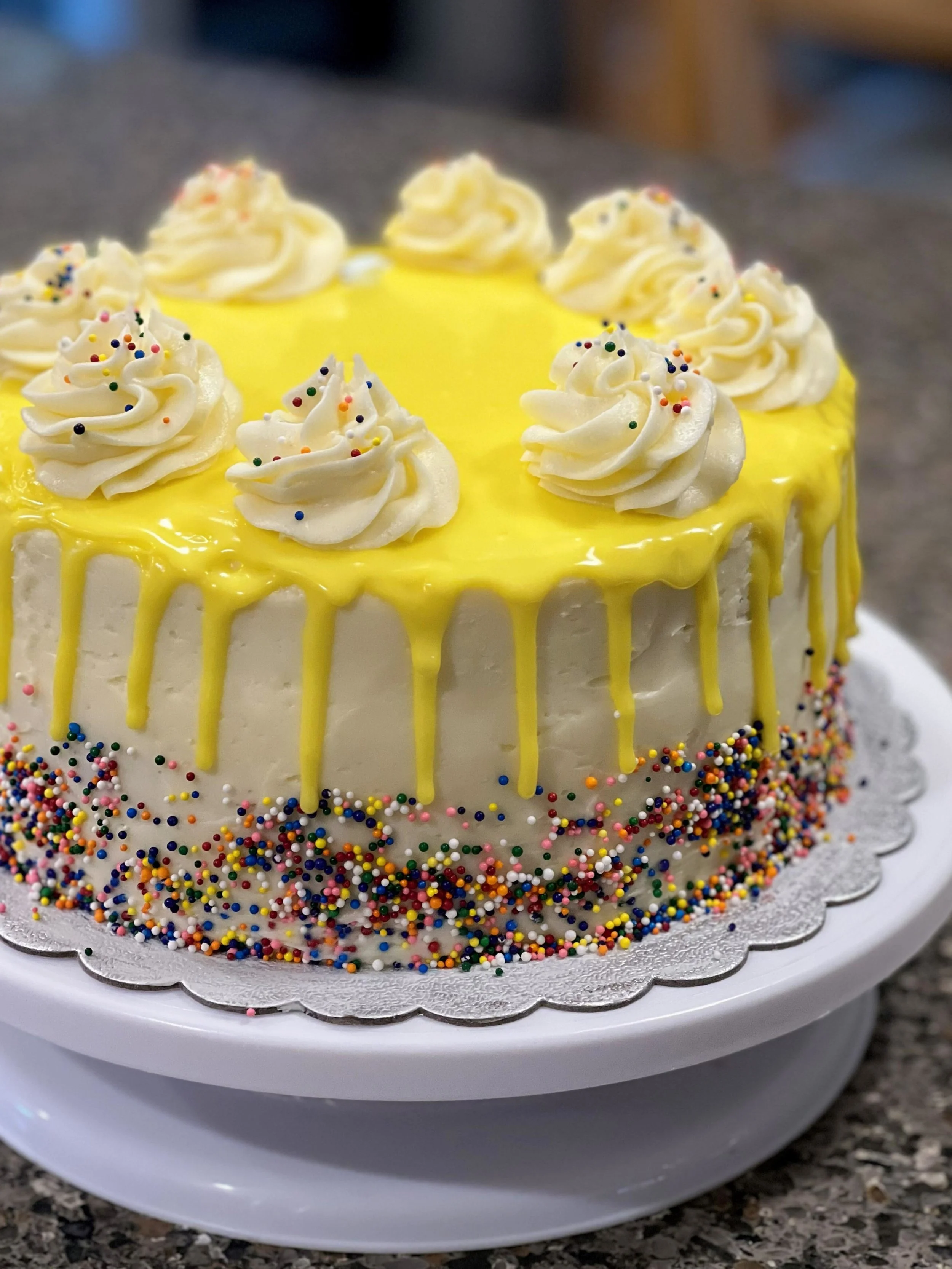 A round cake with yellow drip icing, decorated with white frosting swirls and colorful sprinkles around the base, placed on a white cake stand.