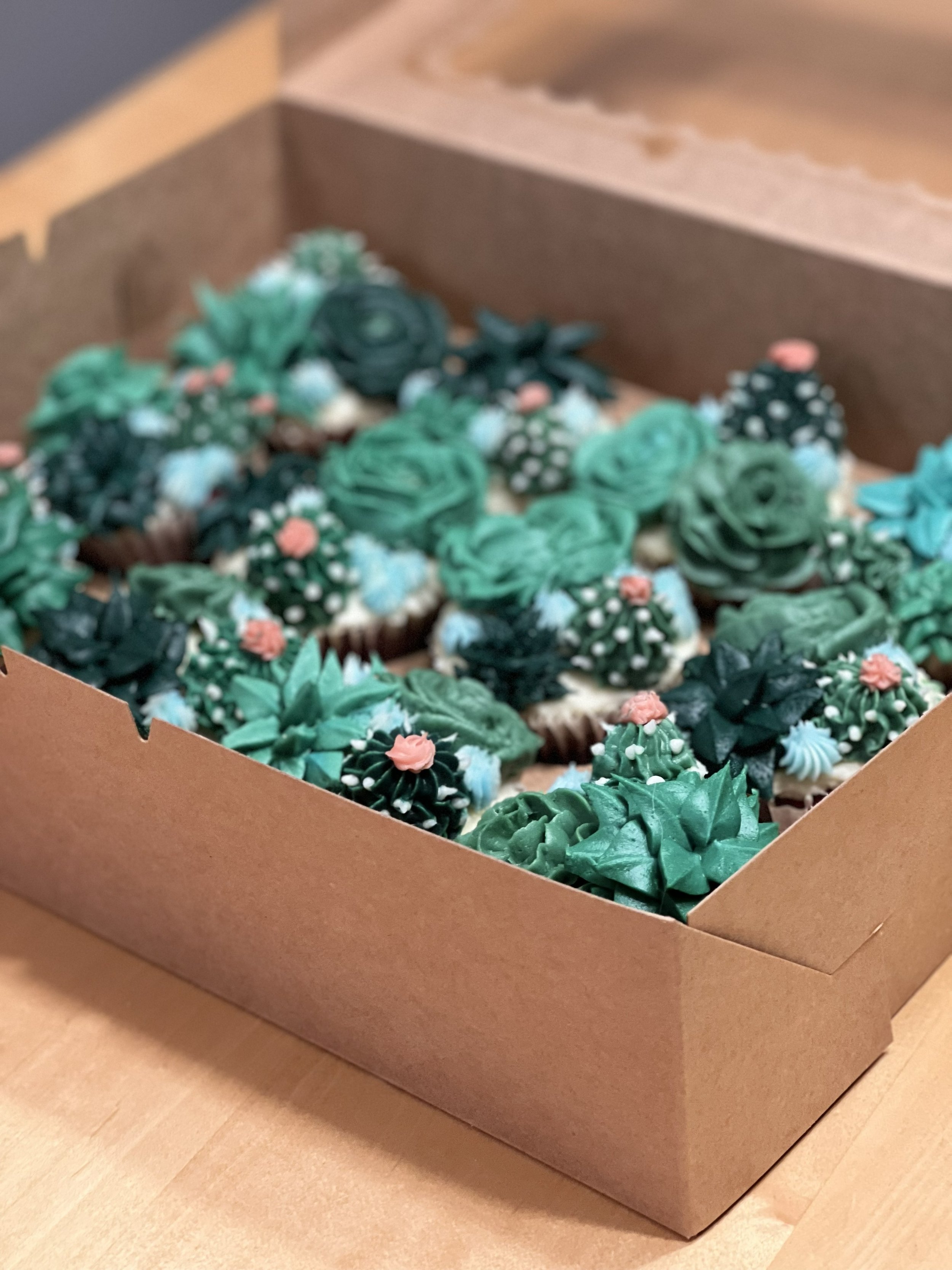 A cardboard box filled with decorated cupcakes featuring green, blue, and pink frosting flowers and designs.