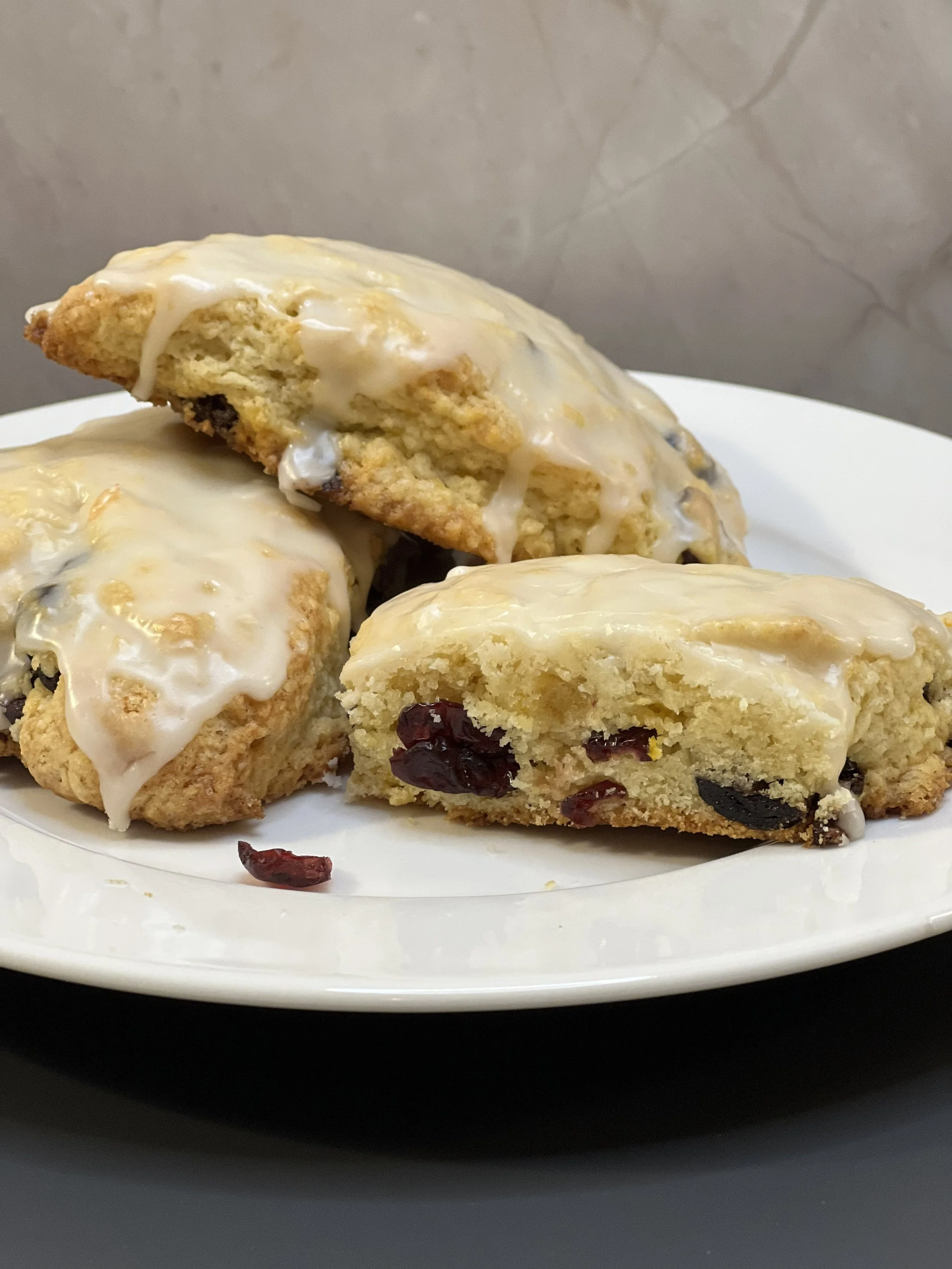 Three cranberry cookies with white icing on a white plate, one is broken in half showing cranberries inside.