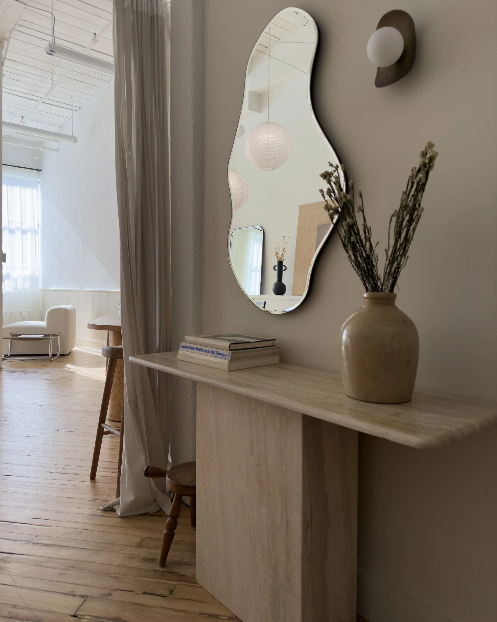 A beige marble console table with books and a vase of dried flowers, a wavy mirror on the wall, and a wall-mounted light fixture, with a glimpse of a room with white furniture, curtains, and paper lanterns reflected in the mirror.