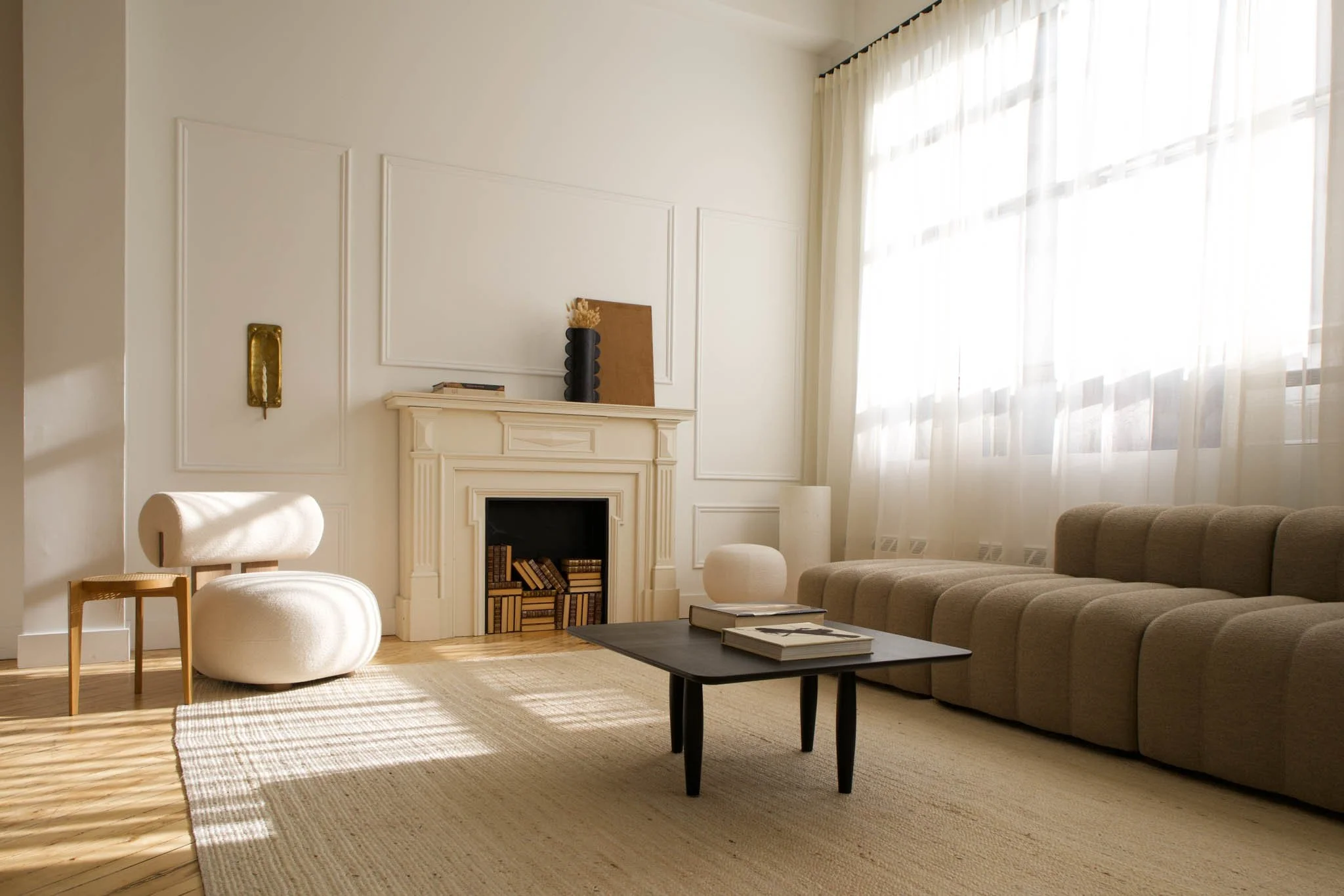Living room with beige sectional sofa, white curtains, black coffee table, and fireplace with books, decorated with vases and artwork.