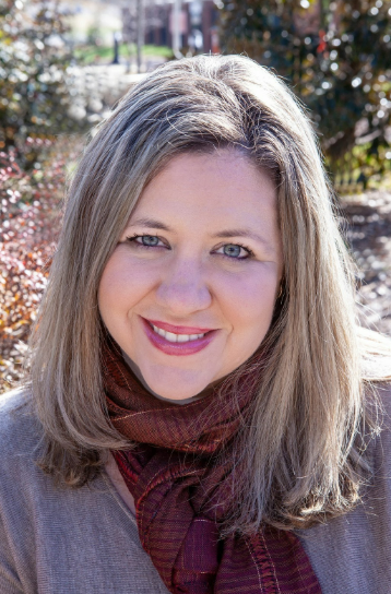 A woman with shoulder-length blonde hair smiling outdoors with trees and a stone wall in the background.