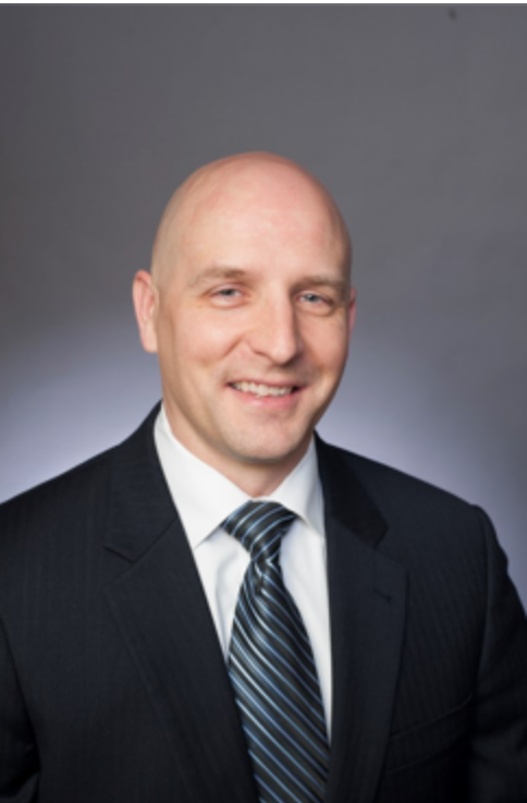 A professional man wearing a black suit, striped tie, and white shirt, smiling at the camera against a gray background.