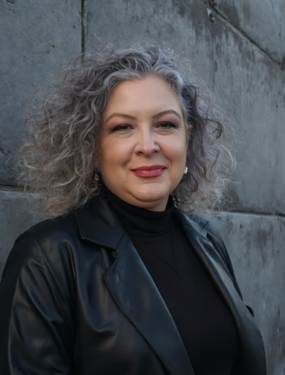 A woman with curly gray hair wearing a black leather jacket and black turtleneck, standing against a stone wall.