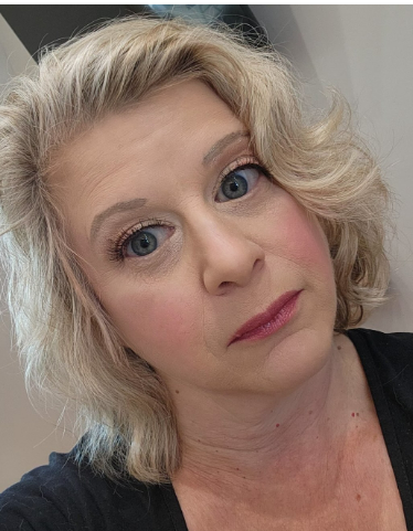 A close-up of a woman with blonde curly hair, blue eyes, and wearing makeup, looking at the camera.