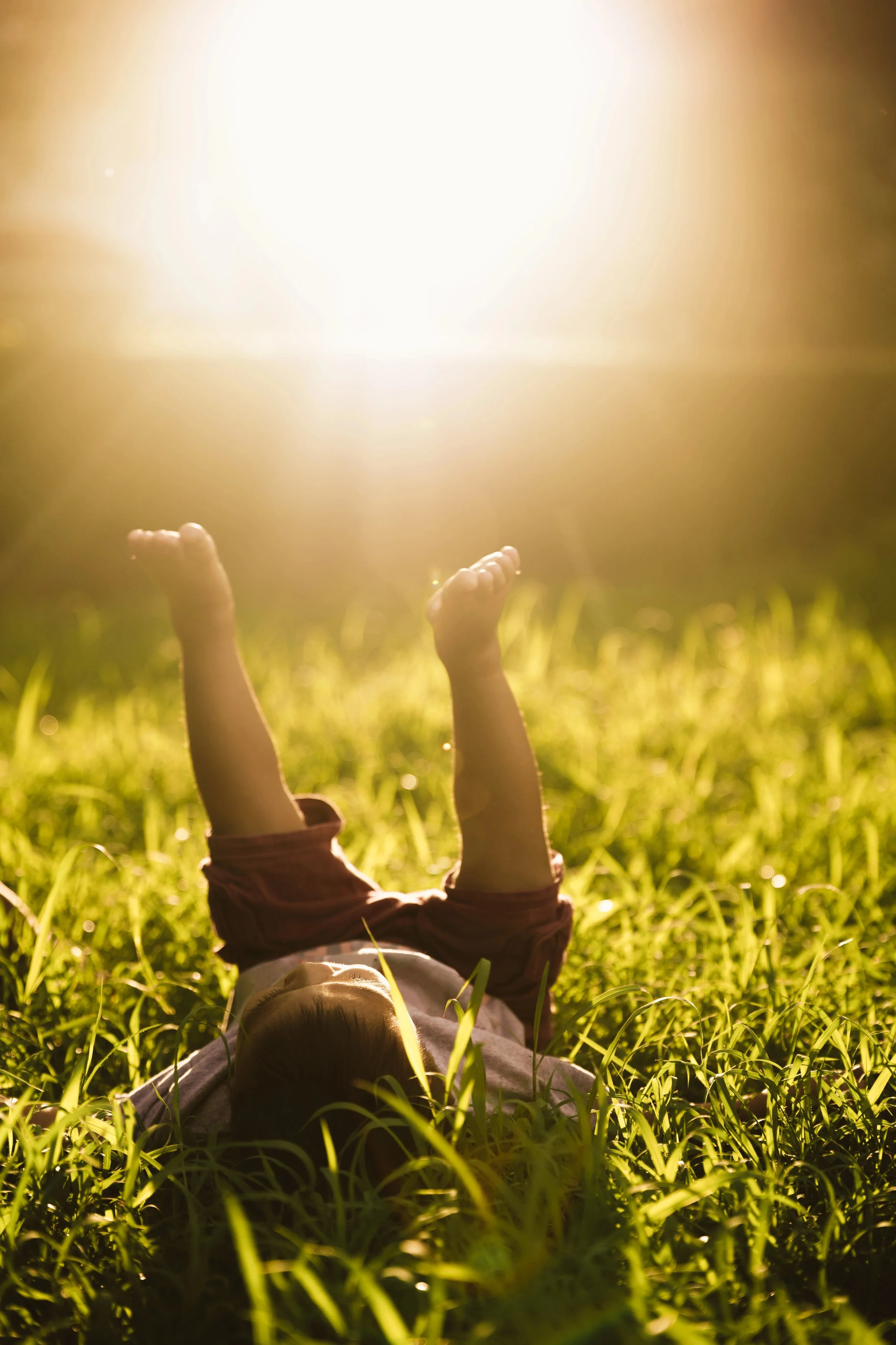 Child lying on grass with arms raised towards bright sunlight.