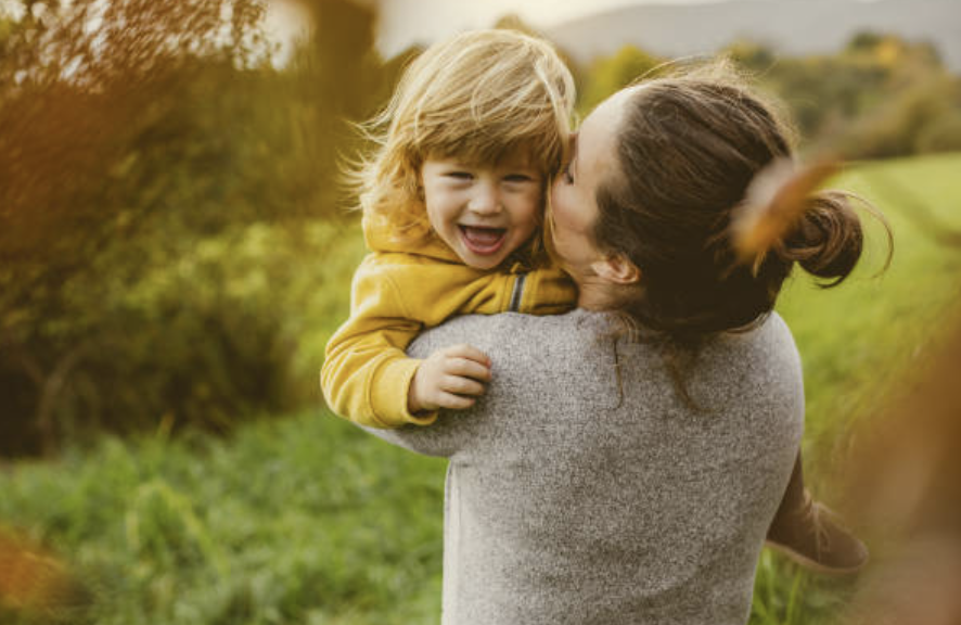 A woman holding a laughing young child outside in a grassy area during daytime.
