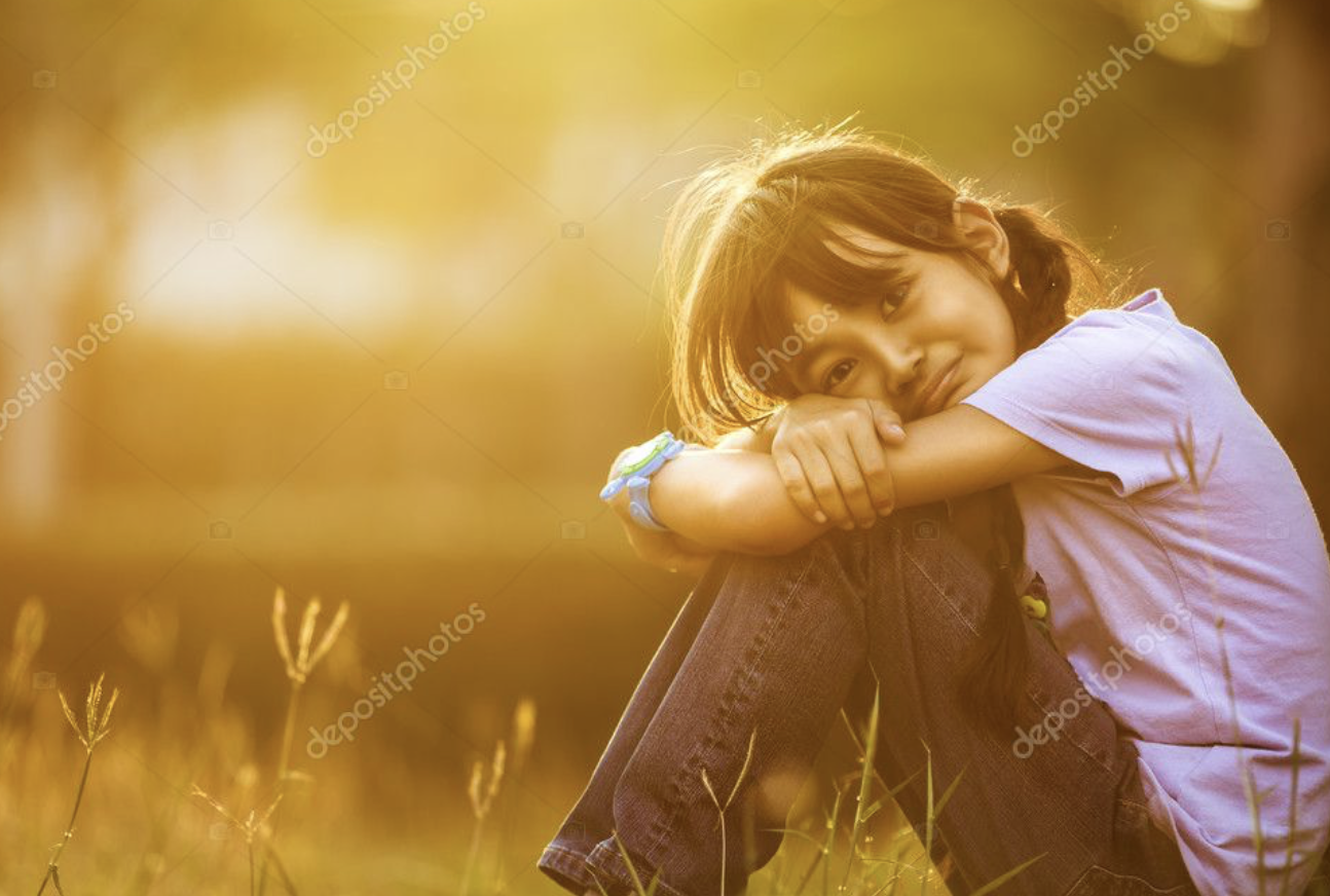 A young girl sitting outdoors during sunset with her arms crossed on his knees, smiling softly at the camera.