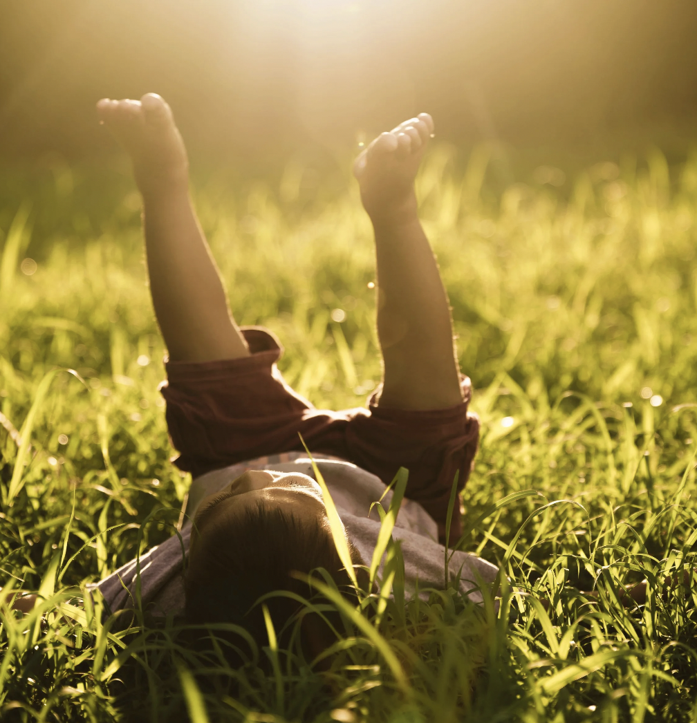 Happy child lying in a grassy field, arms raised towards the sky, during sunset or sunrise with warm, golden light.