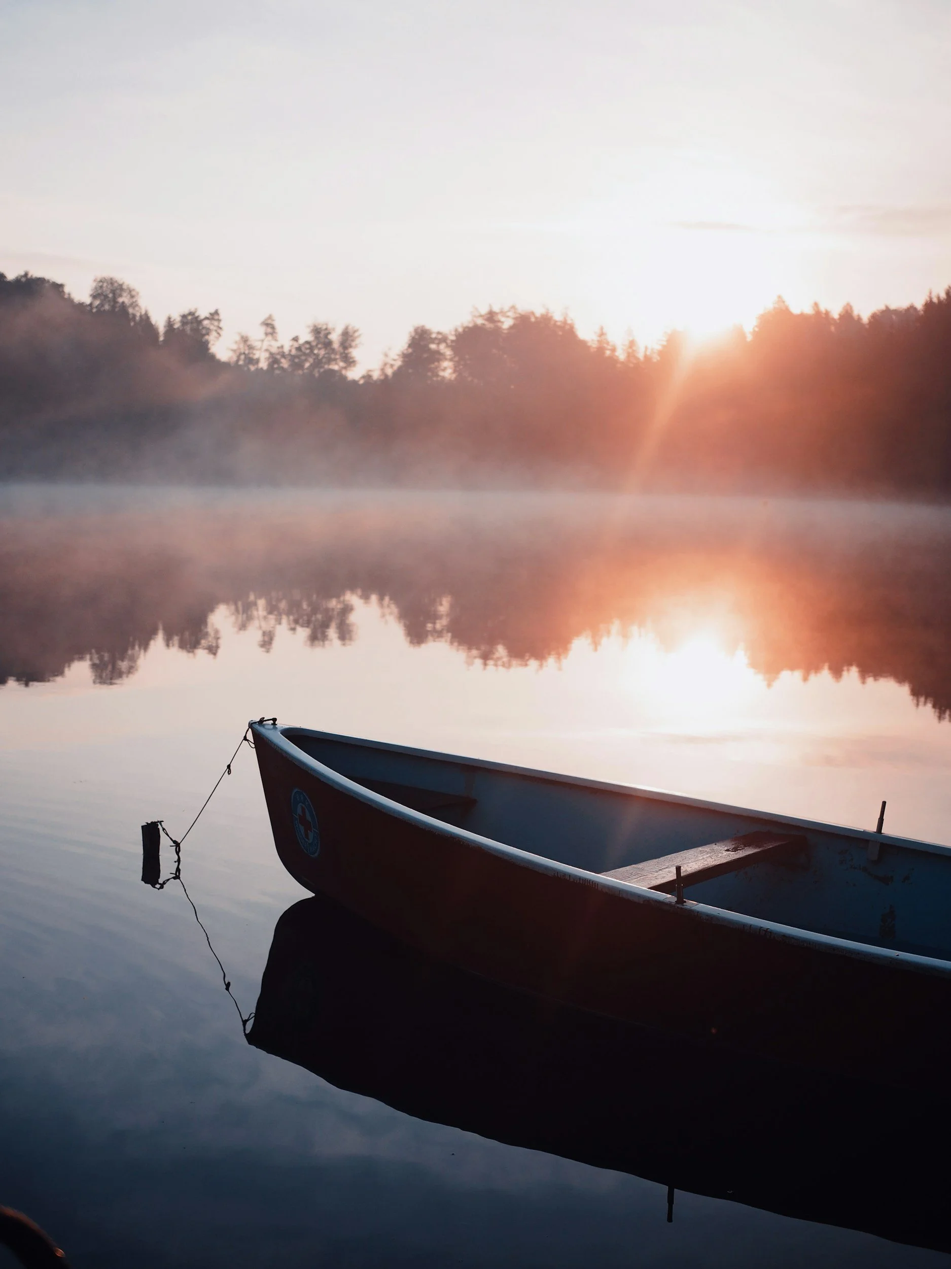 A boat floating on a calm lake during sunrise or sunset, with mist rising over the water and trees visible in the background.