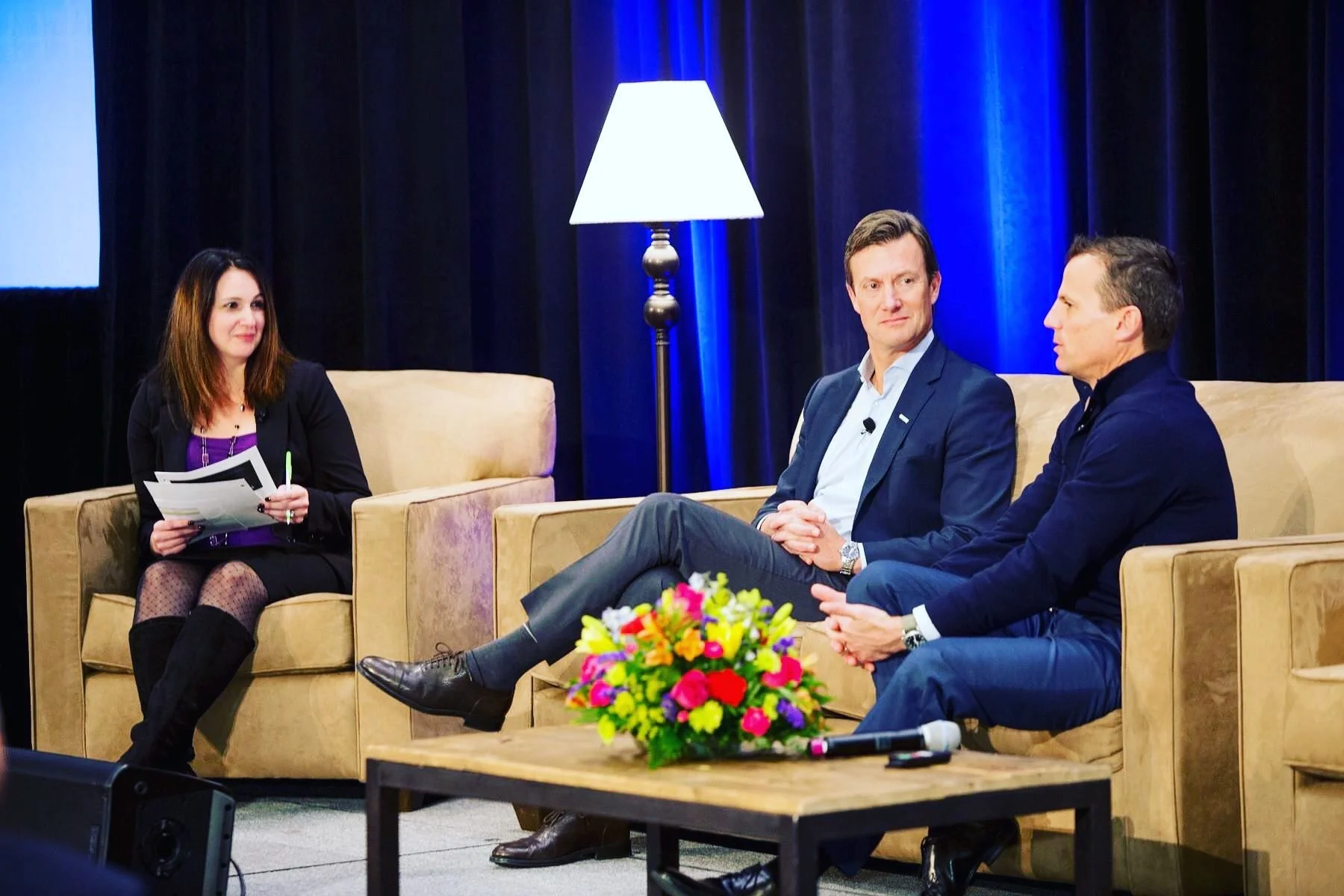 A woman in a black blazer and purple top sits on a beige sofa, holding papers and a pen. Two men in dark suits sit on nearby beige sofas, engaged in conversation. There is a colorful flower arrangement on a table in the foreground, and a blue-lit curtain with a tall lamp behind them.