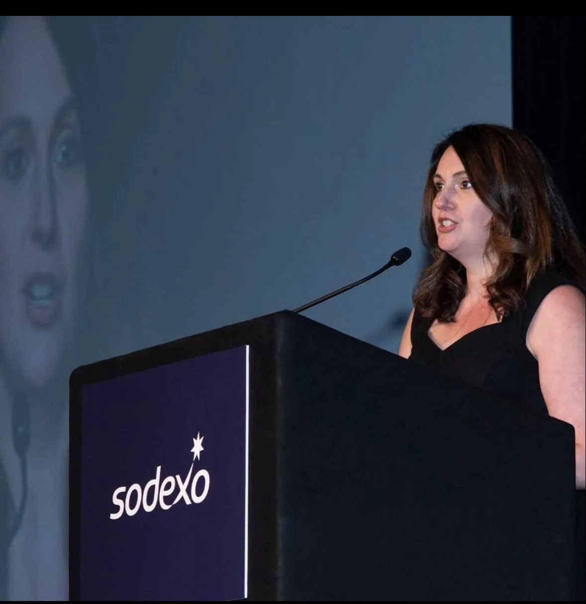 A woman with dark brown hair speaking at a podium with a sodexo logo, with a large screen behind her displaying her face.