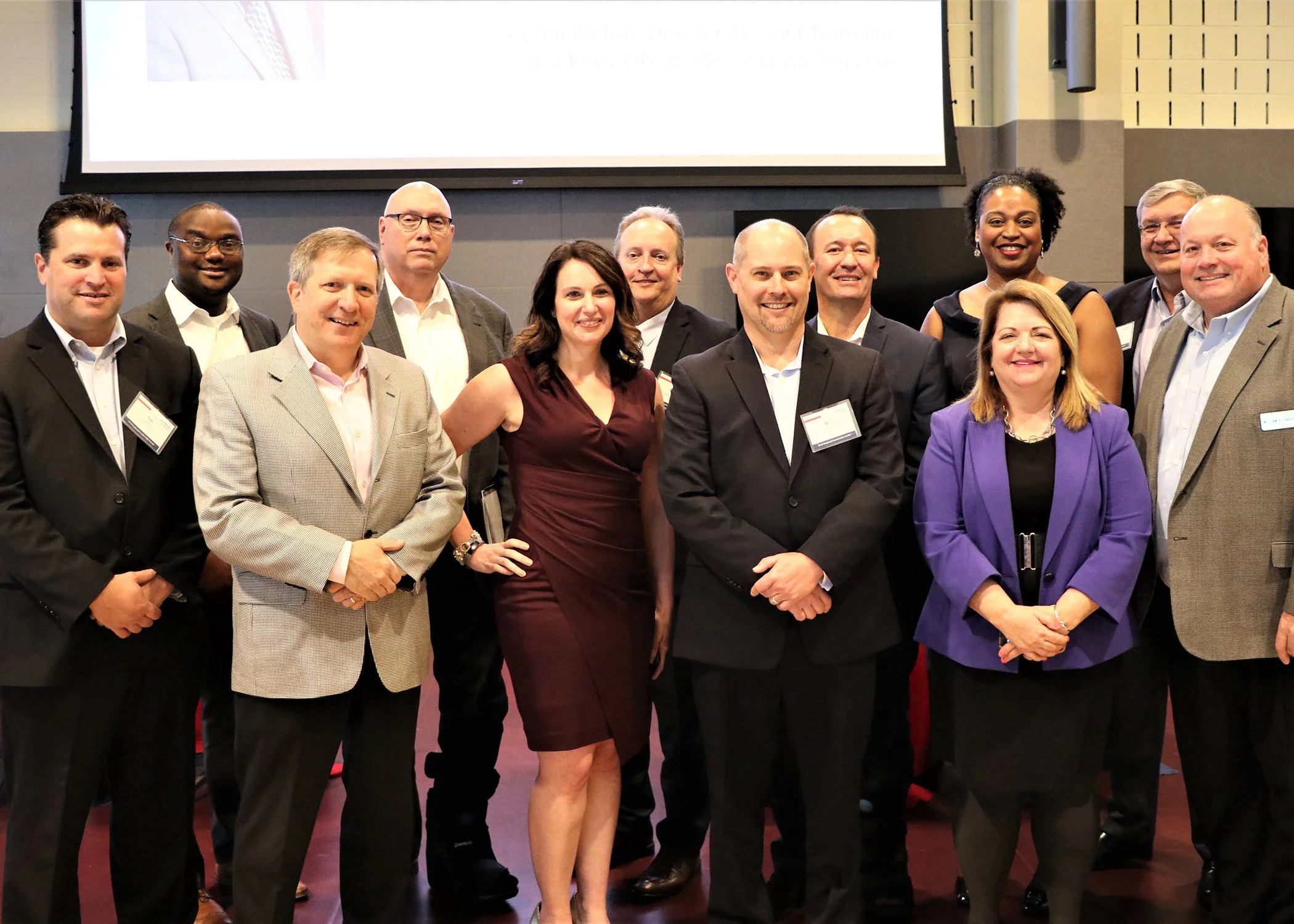 A group of eleven professionally dressed people standing together in a conference room, smiling for a photo with a large presentation screen behind them.