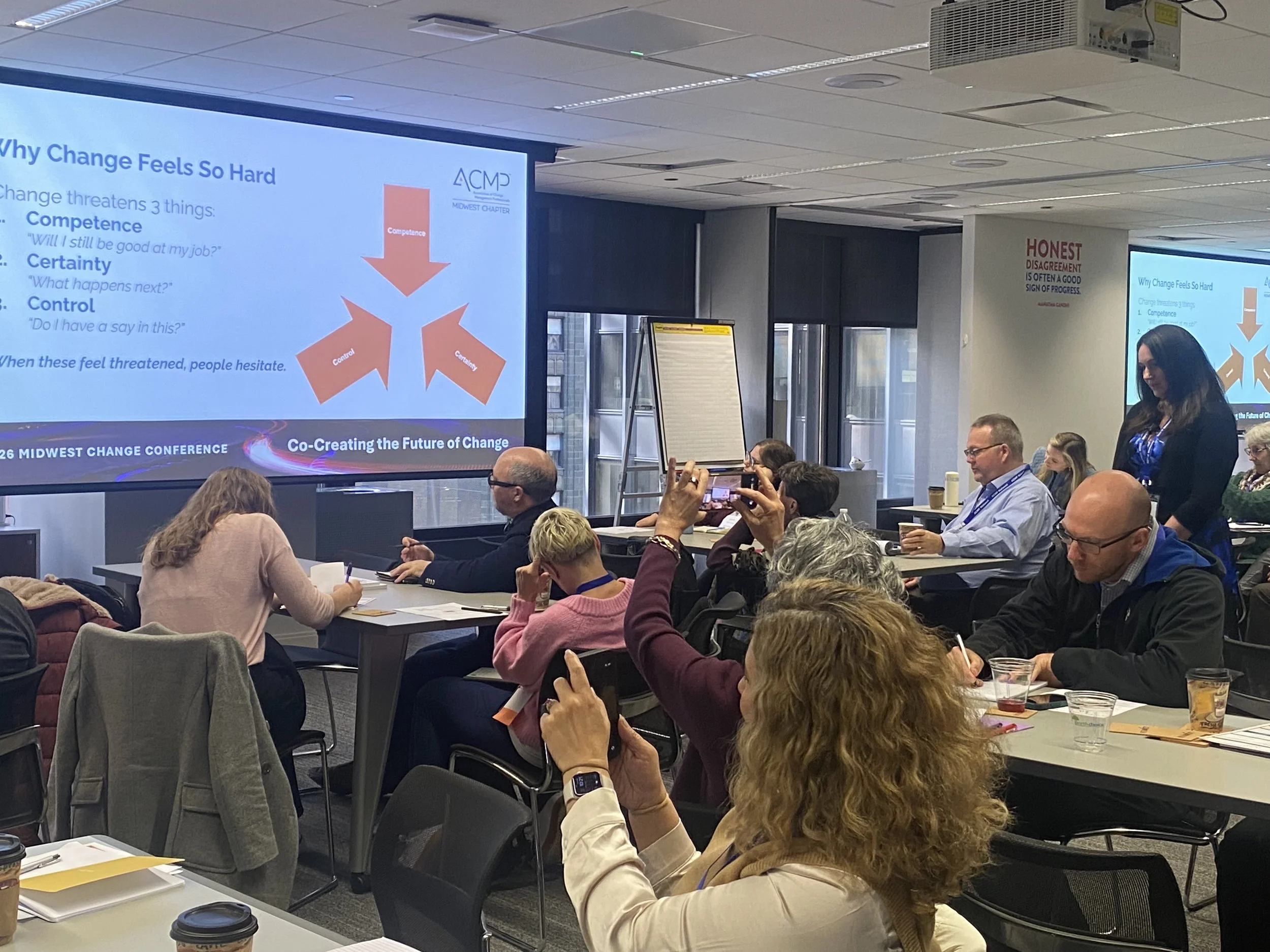 A conference room filled with attendees sitting at tables, some taking notes and others taking photos. A large screen displays a PowerPoint slide titled 'Why Change Feels So Hard' with bullet points about change threats. The room has a modern design with large windows and ceiling lights.