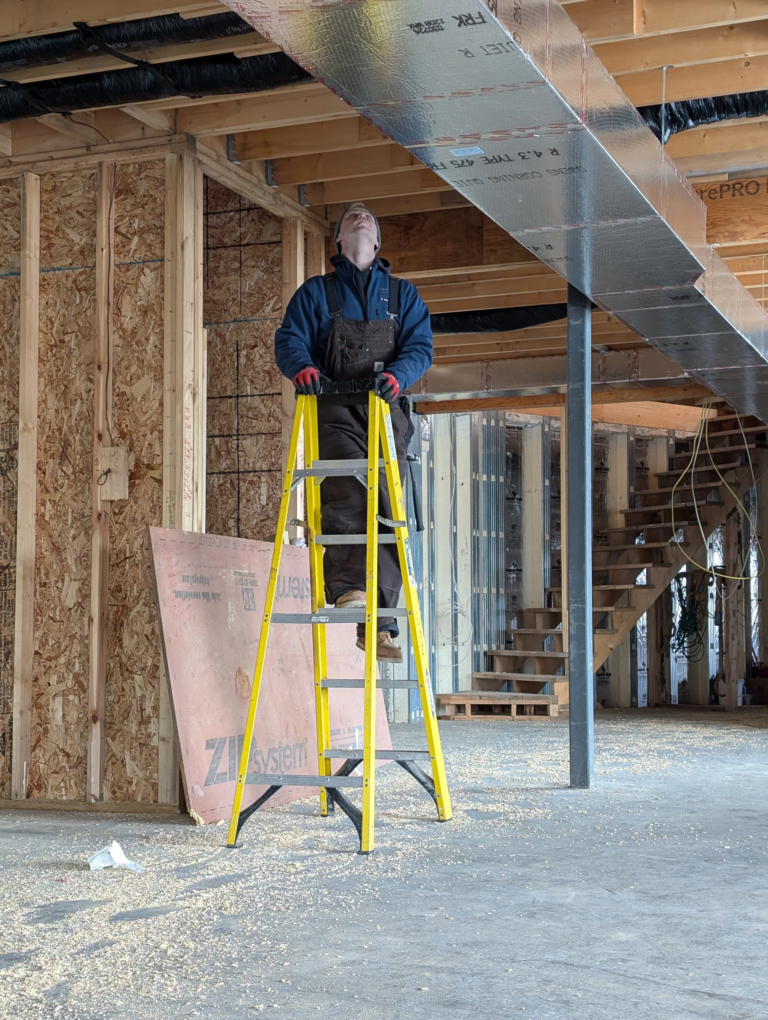 Construction worker looking up at ceiling structure under construction, standing on a yellow ladder in a partially built building with exposed wooden framework and insulation materials.