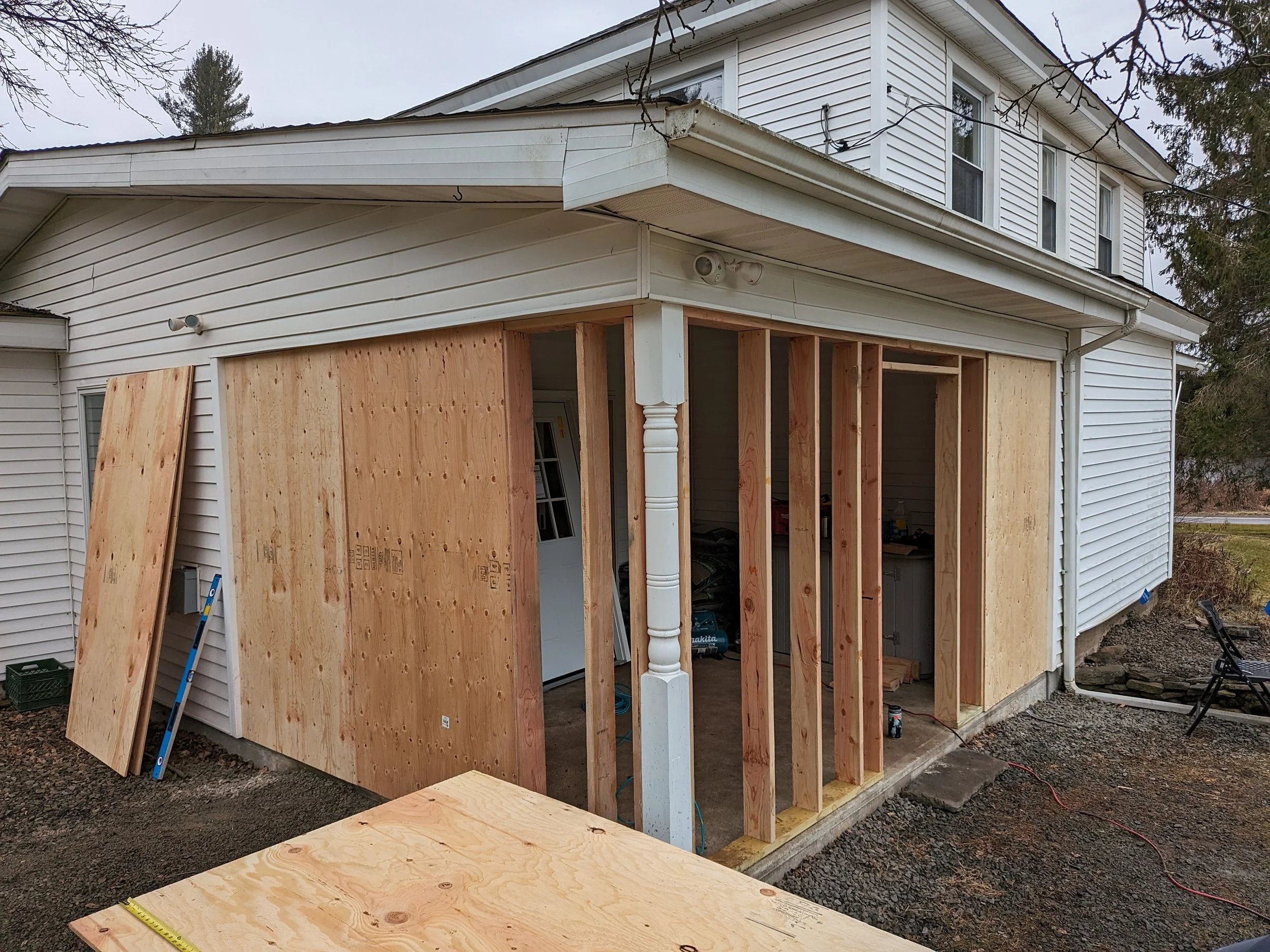 Side view of a house under construction with plywood panels on the exterior wall, wooden framing, and white vinyl siding on the upper level.