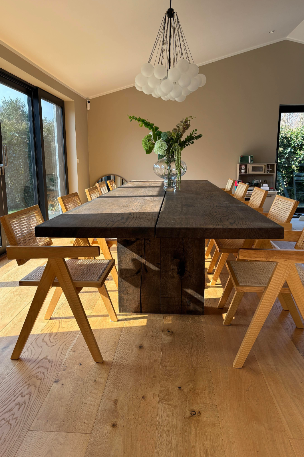 Dining room with a large wooden table and ten wooden chairs, a glass vase with green flowers, modern chandelier, large sliding glass door, and wooden floor.