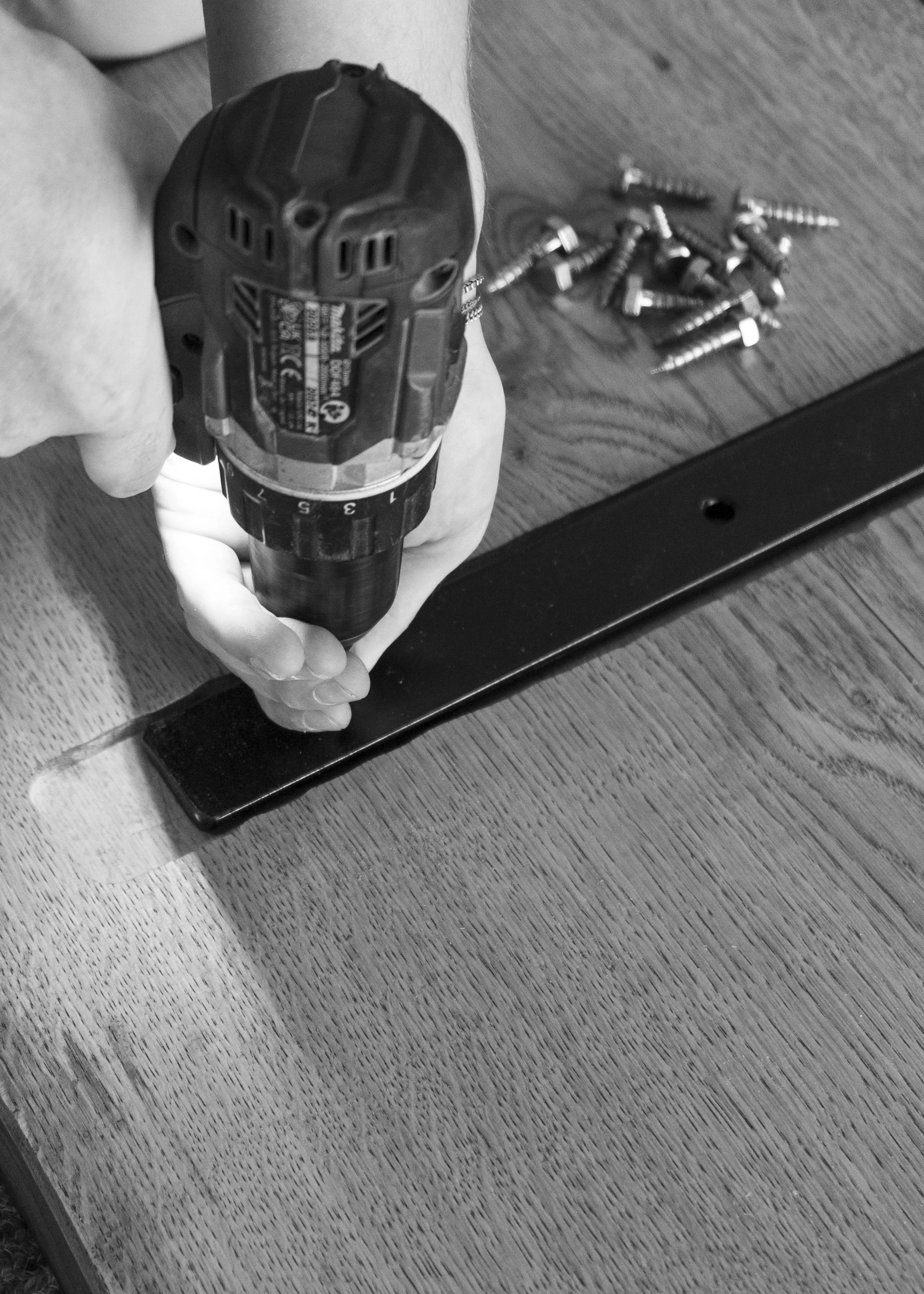 Person using a cordless drill on a piece of wood, with screws scattered on the surface.