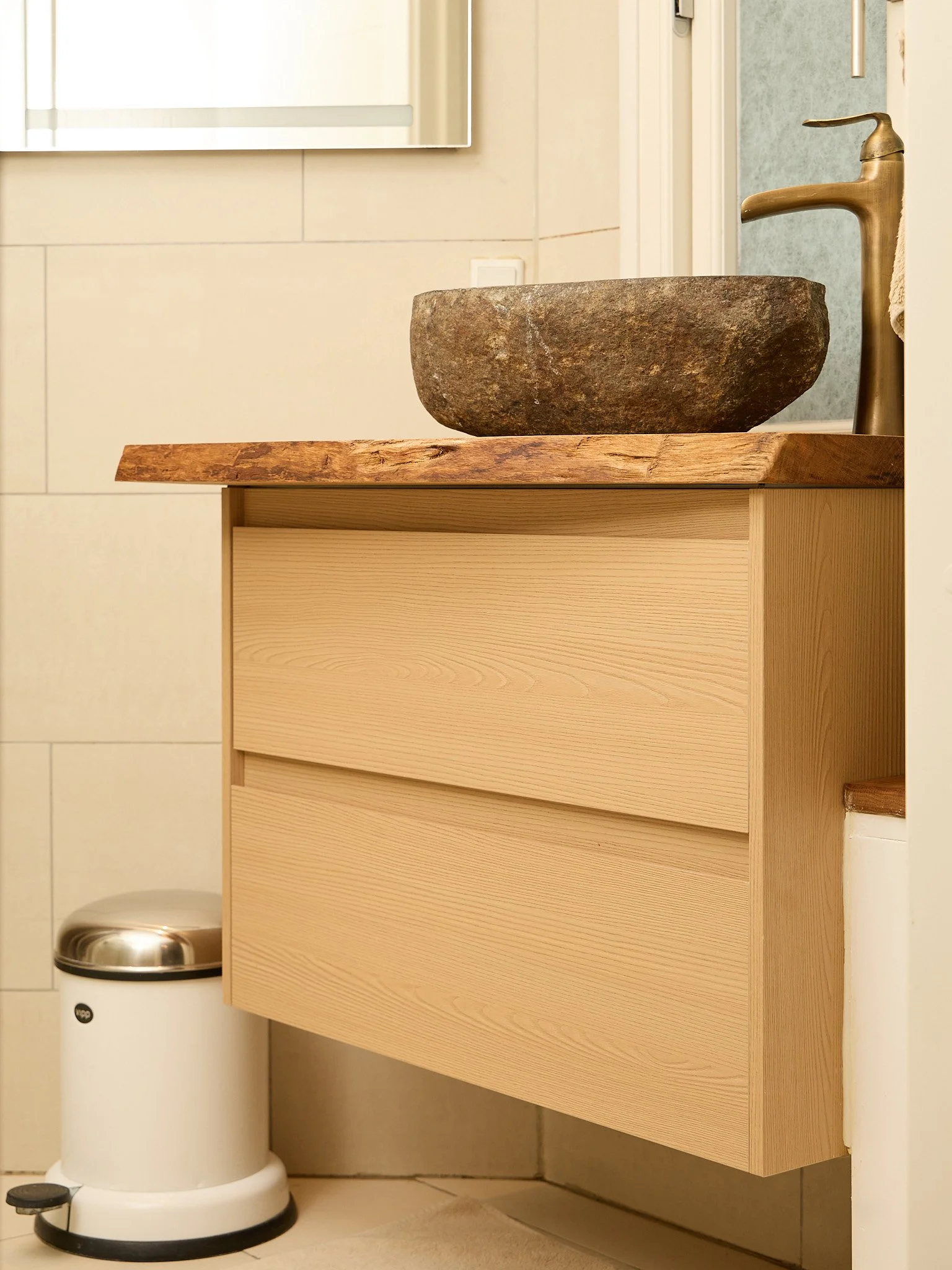 A bathroom sink with a stone basin on a wooden counter, next to a faucet and a small trash bin.