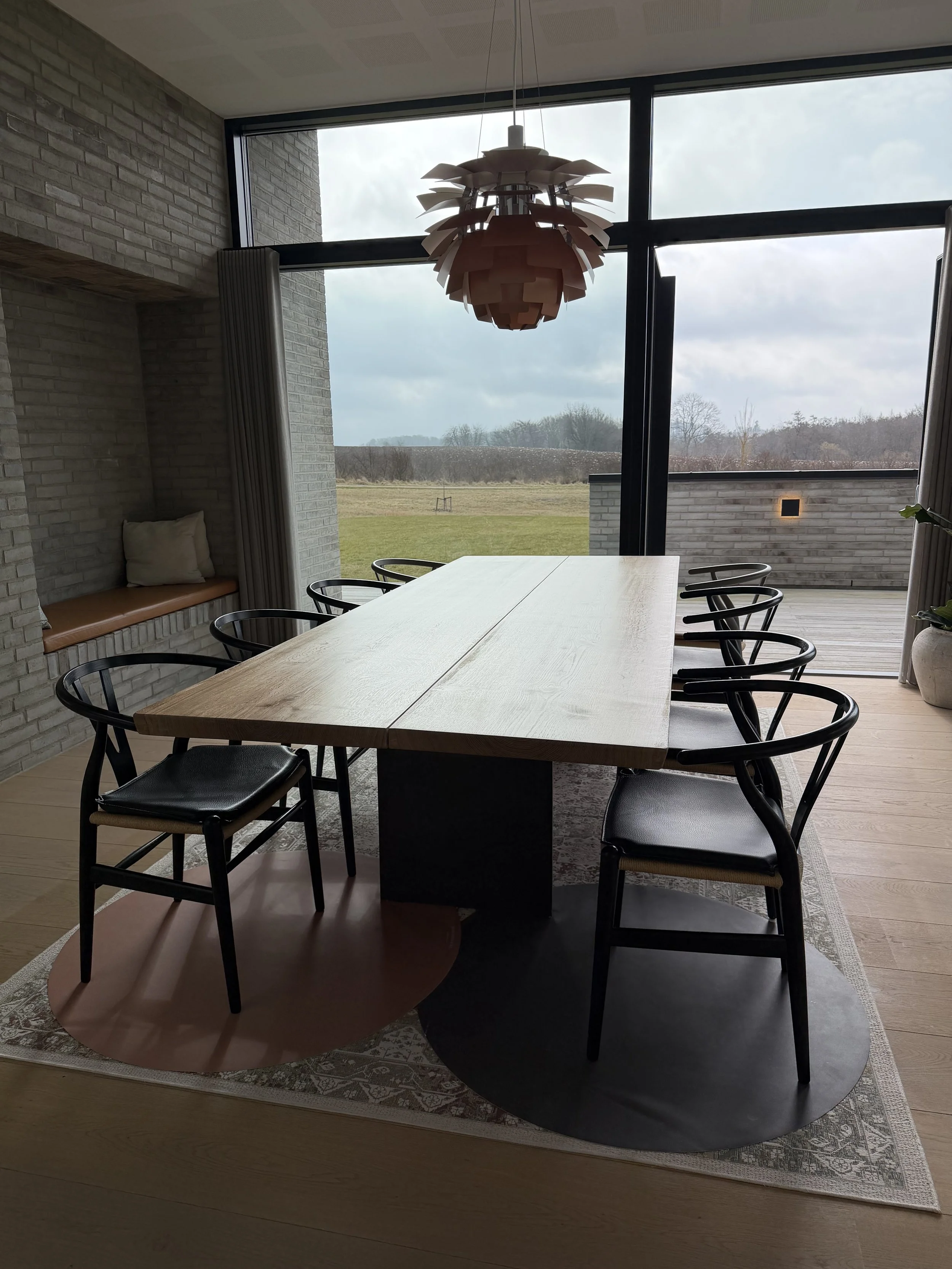 Interior of a modern dining room with a large wooden table, black chairs, a contemporary pendant light, large windows offering a view of a grassy field and cloudy sky outside, and a built-in window seat with cushion on the left side.