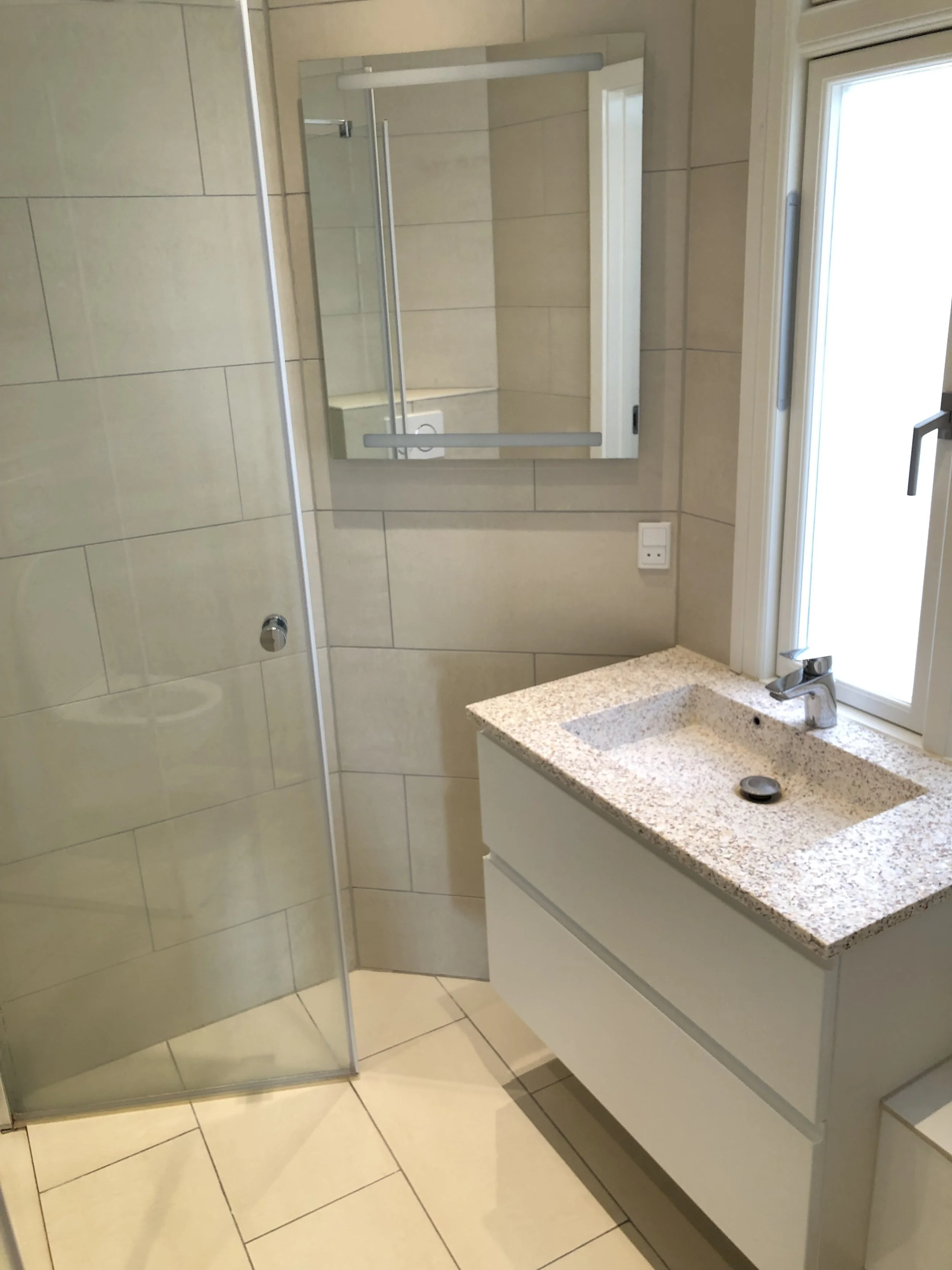 A modern bathroom with beige tiled walls, a mirror, a white vanity with a granite countertop, a sink, and a window. There is a glass shower enclosure and a towel rail visible.