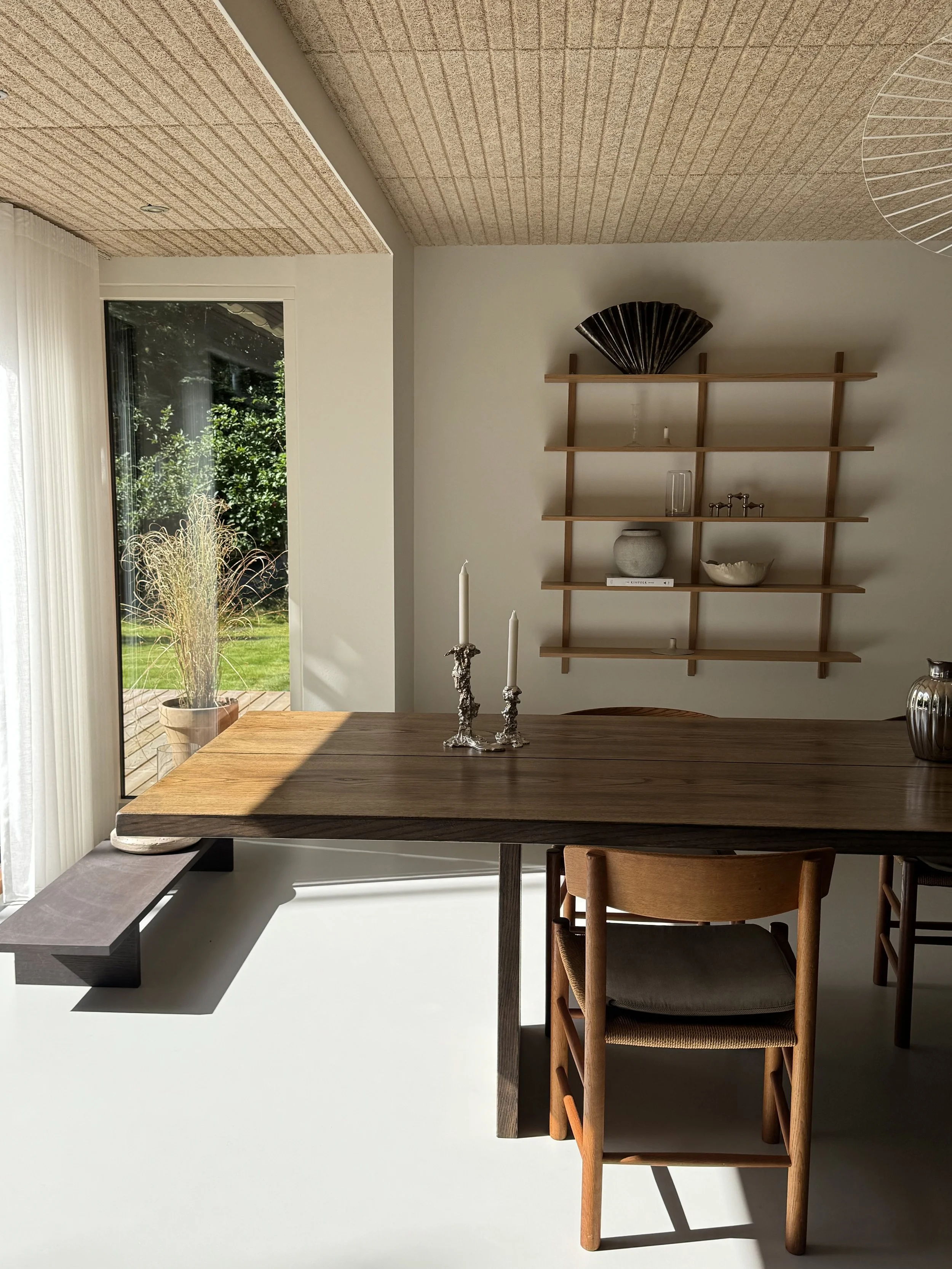 Modern dining room with a large wooden table, a wooden chair with a cushion, a wall-mounted wooden shelf with decorative objects, a large window with sheer white curtains, and a view of green outdoor plants.