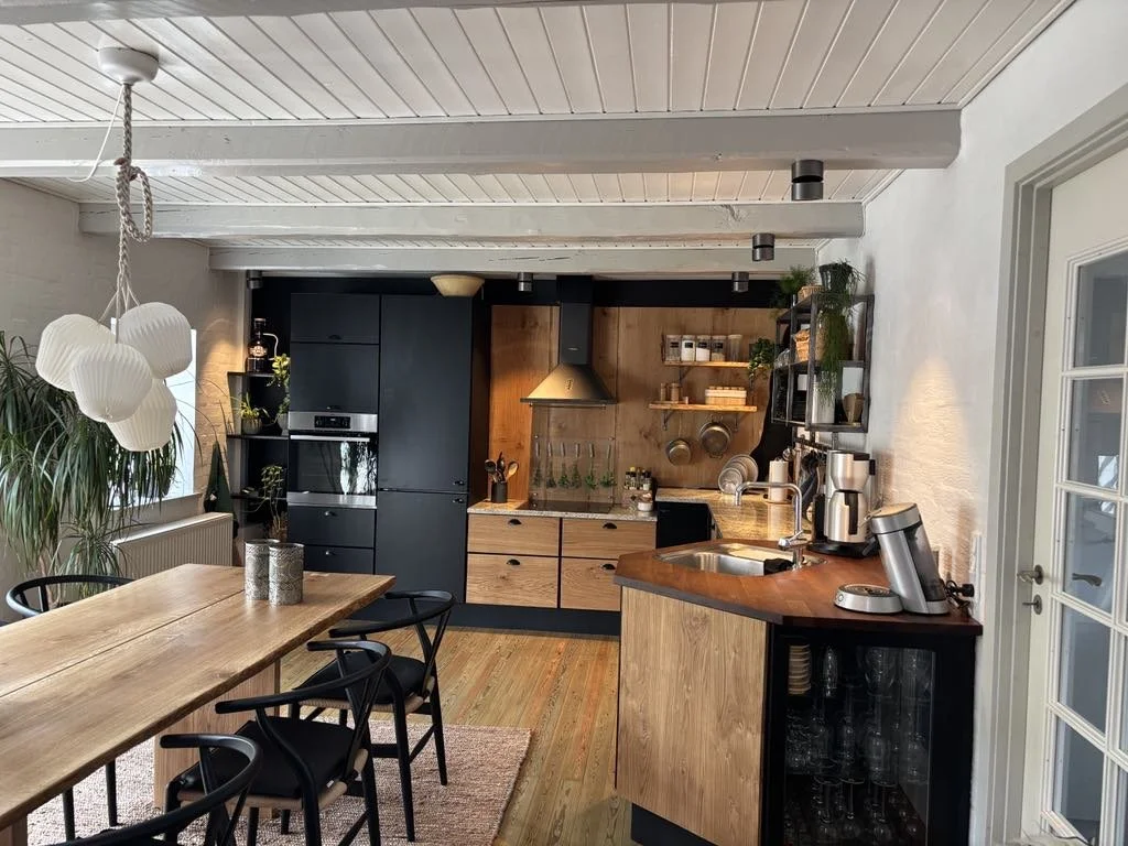 Modern kitchen with black and wooden cabinets, a wooden dining table with black chairs, a hanging white pendant light, and various kitchen appliances. There are plants and open shelves with dishes and jars.