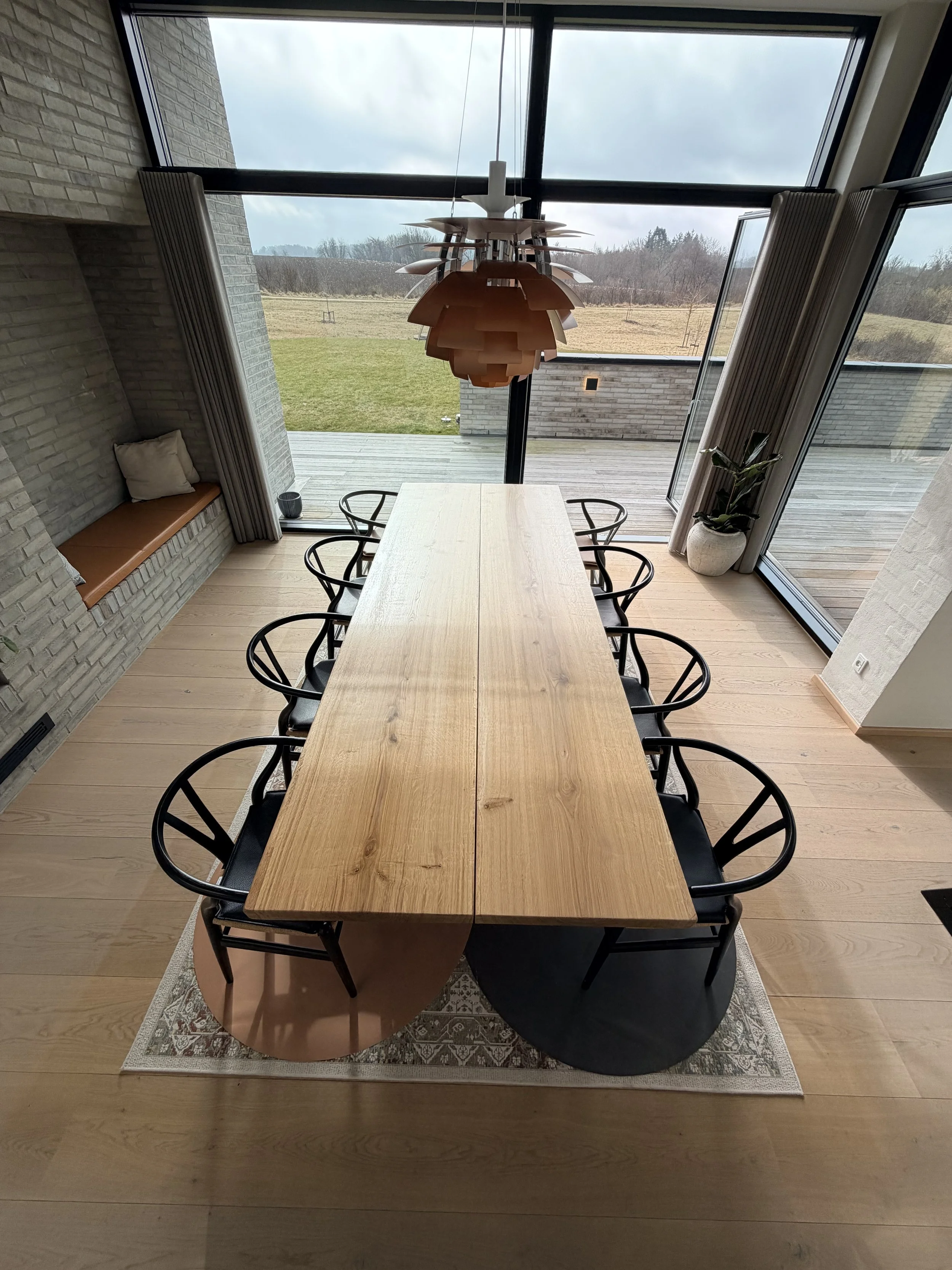 Modern dining room with a long wooden table, black chairs, a potted plant, large windows, and a view of an outdoor deck and natural landscape.