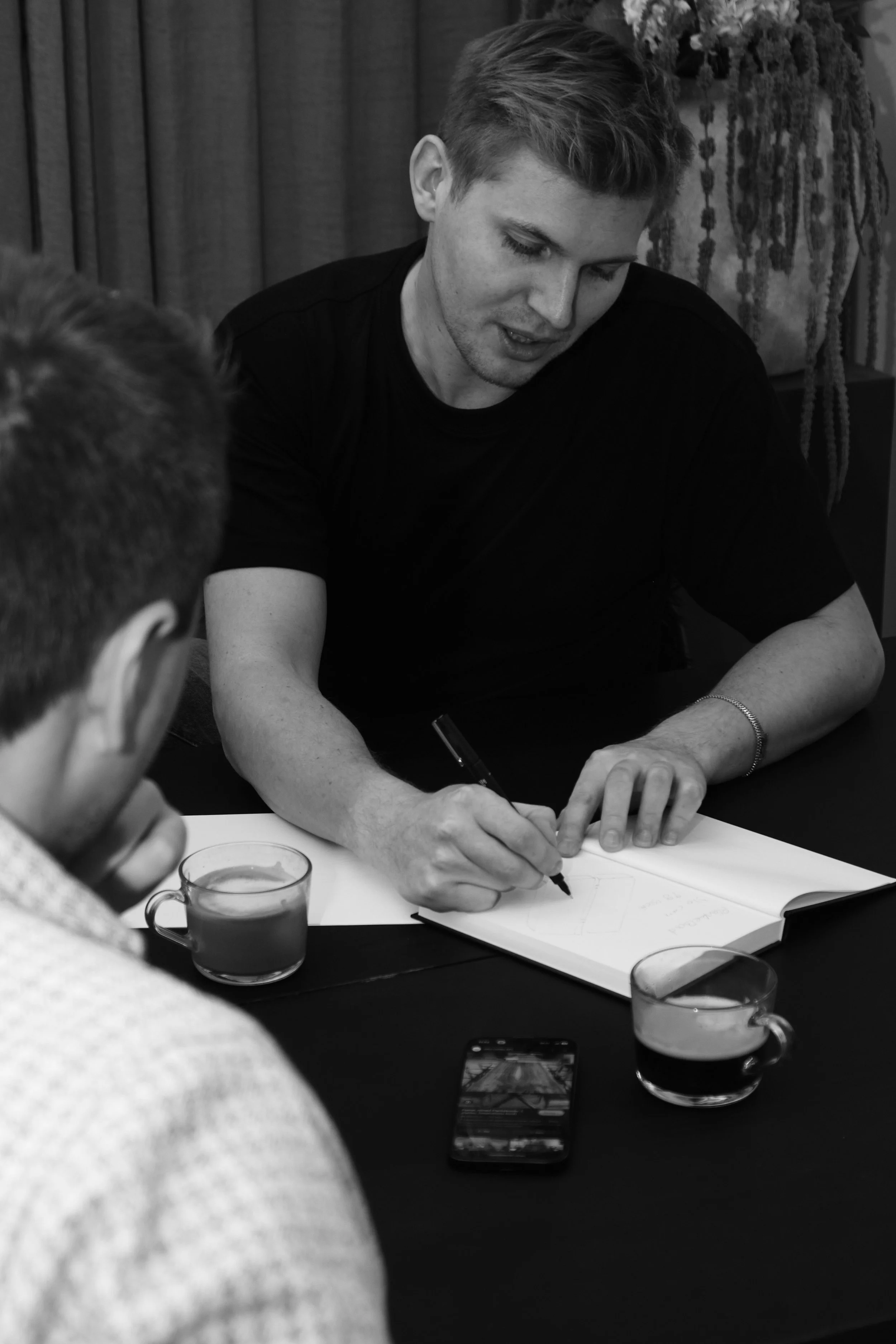 A man with light skin and short hair is signing a book or paper at a table, with a woman with glasses and light hair sitting across from him. There are two cups of coffee or tea and a smartphone on the table. The background has curtains and a large plant.