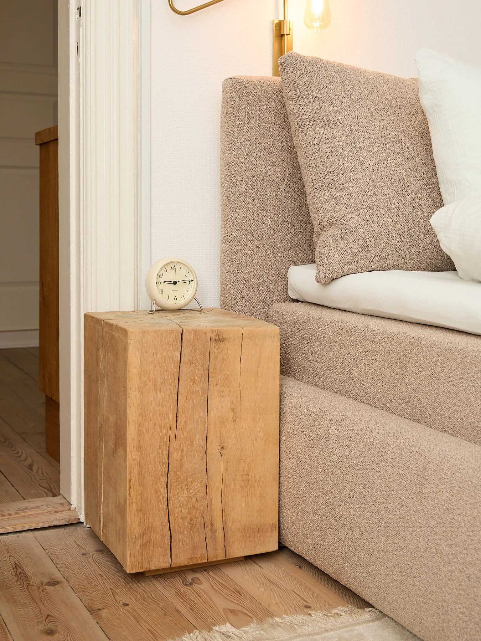 A beige fabric upholstered bed with a matching headboard, white pillows, a wooden nightstand, an analog clock, and a wall-mounted gold lamp with a clear glass bulb in a cozy bedroom.
