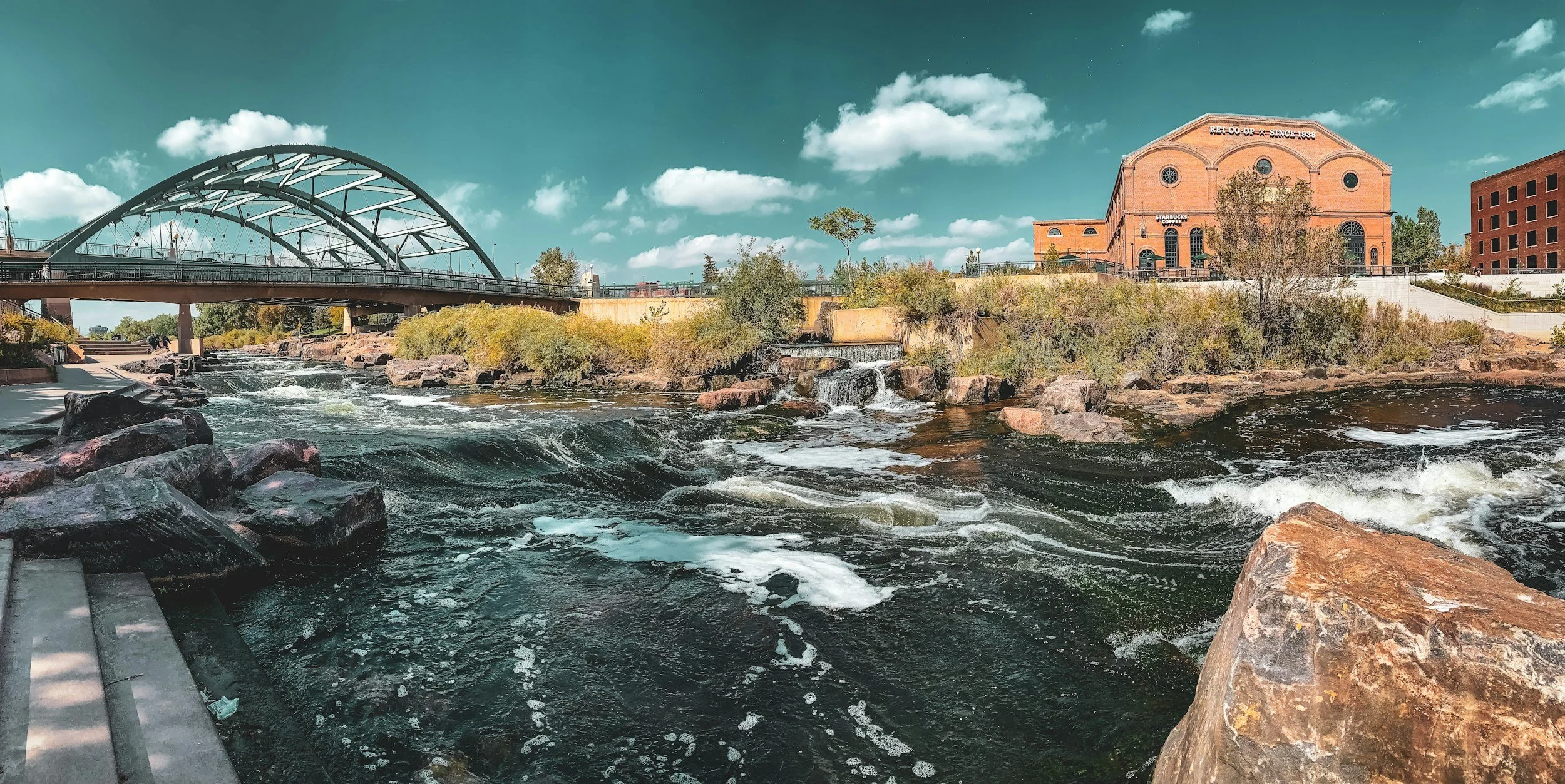 A scenic cityscape featuring a river with rocks and rapids, a bridge, and a brick building with a sign that reads 'REI Co-Op Since 1938' under a partly cloudy sky.