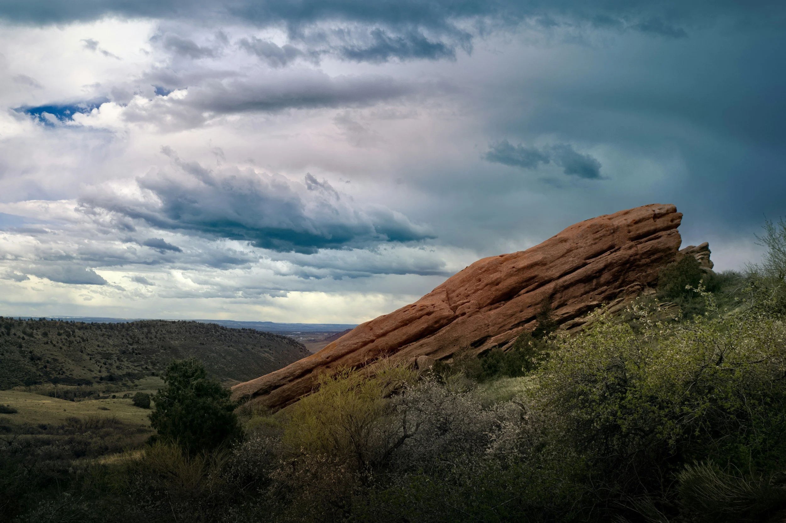 A large, reddish-brown rock formation on a hillside with green shrubs and trees in the foreground and a cloudy sky overhead.