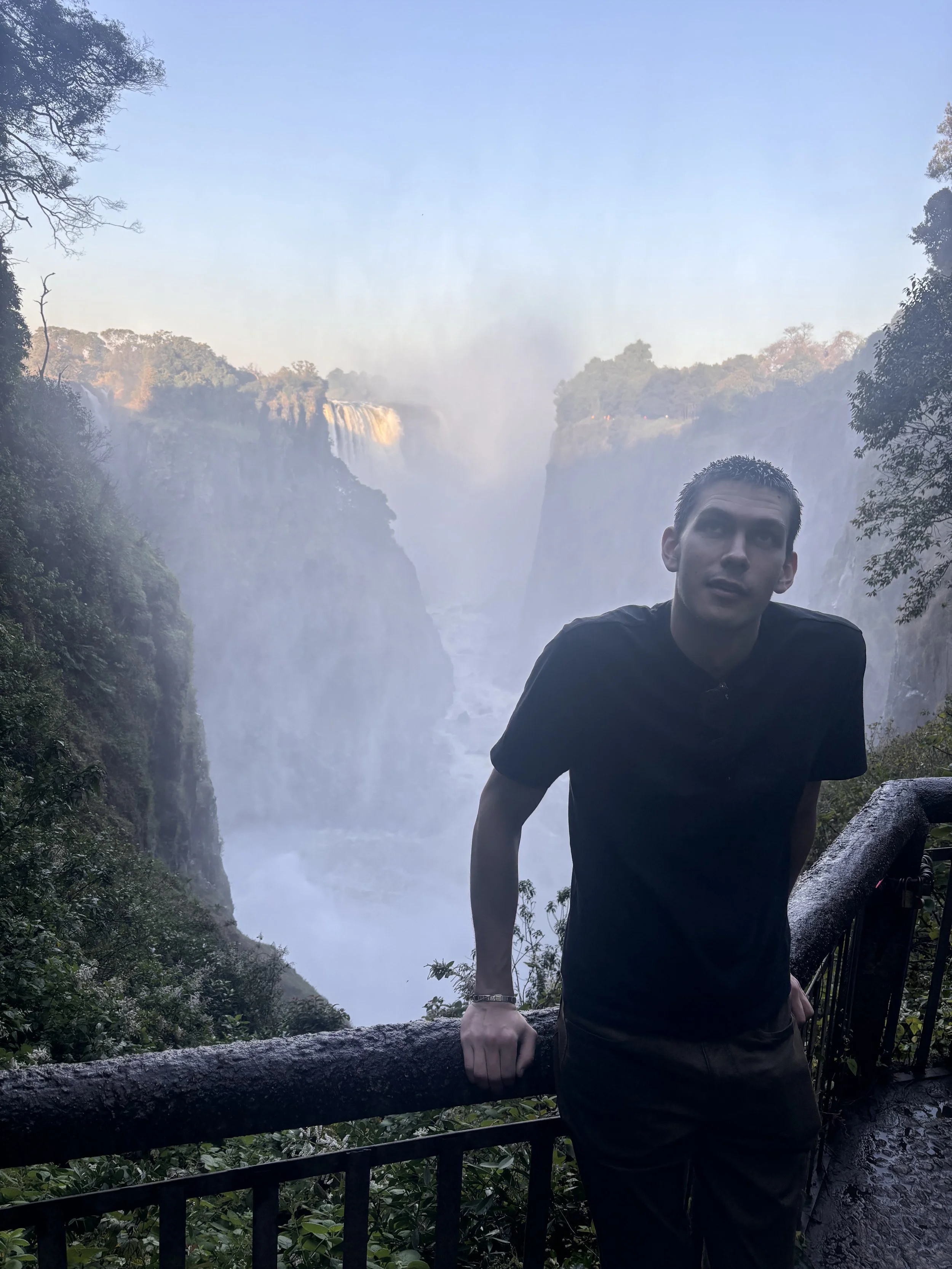 A man leaning on a railing at a scenic waterfall viewpoint in a lush, green forest with mist rising from the waterfall in the background.