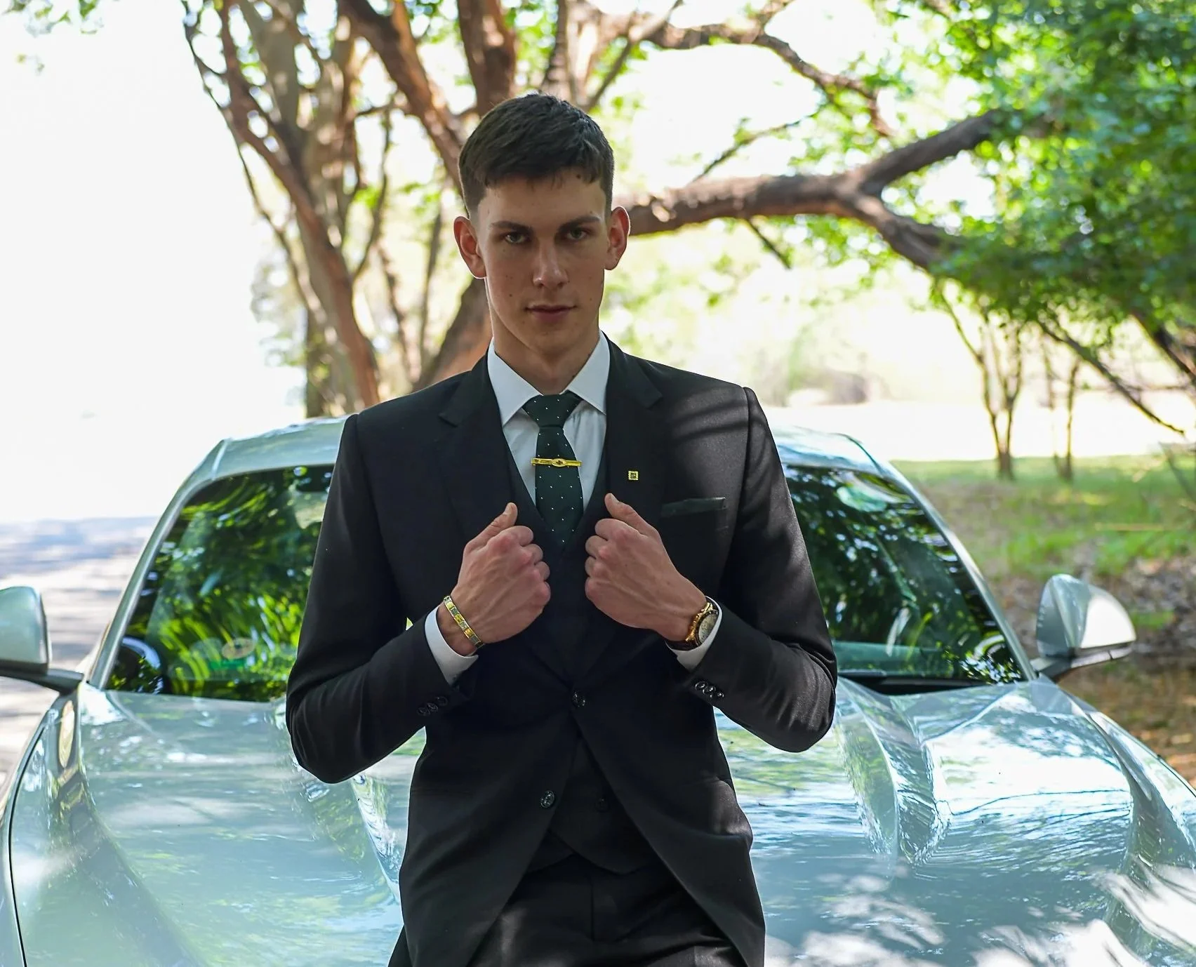 A young man in a black suit and tie stands in front of a silver car with a forested background.
