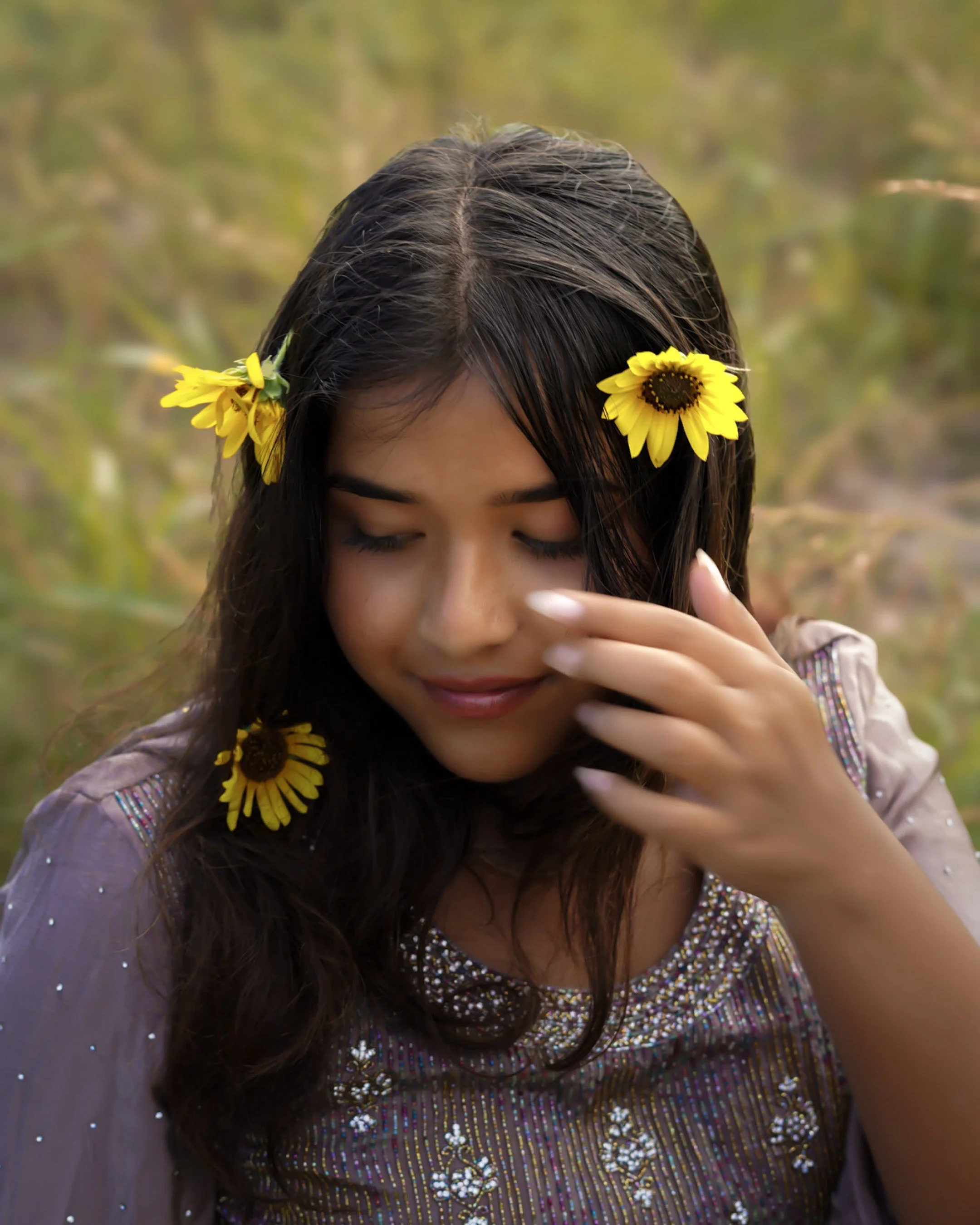 A young woman with dark hair and sunflower accessories in her hair smiling with her eyes closed in an outdoor setting.