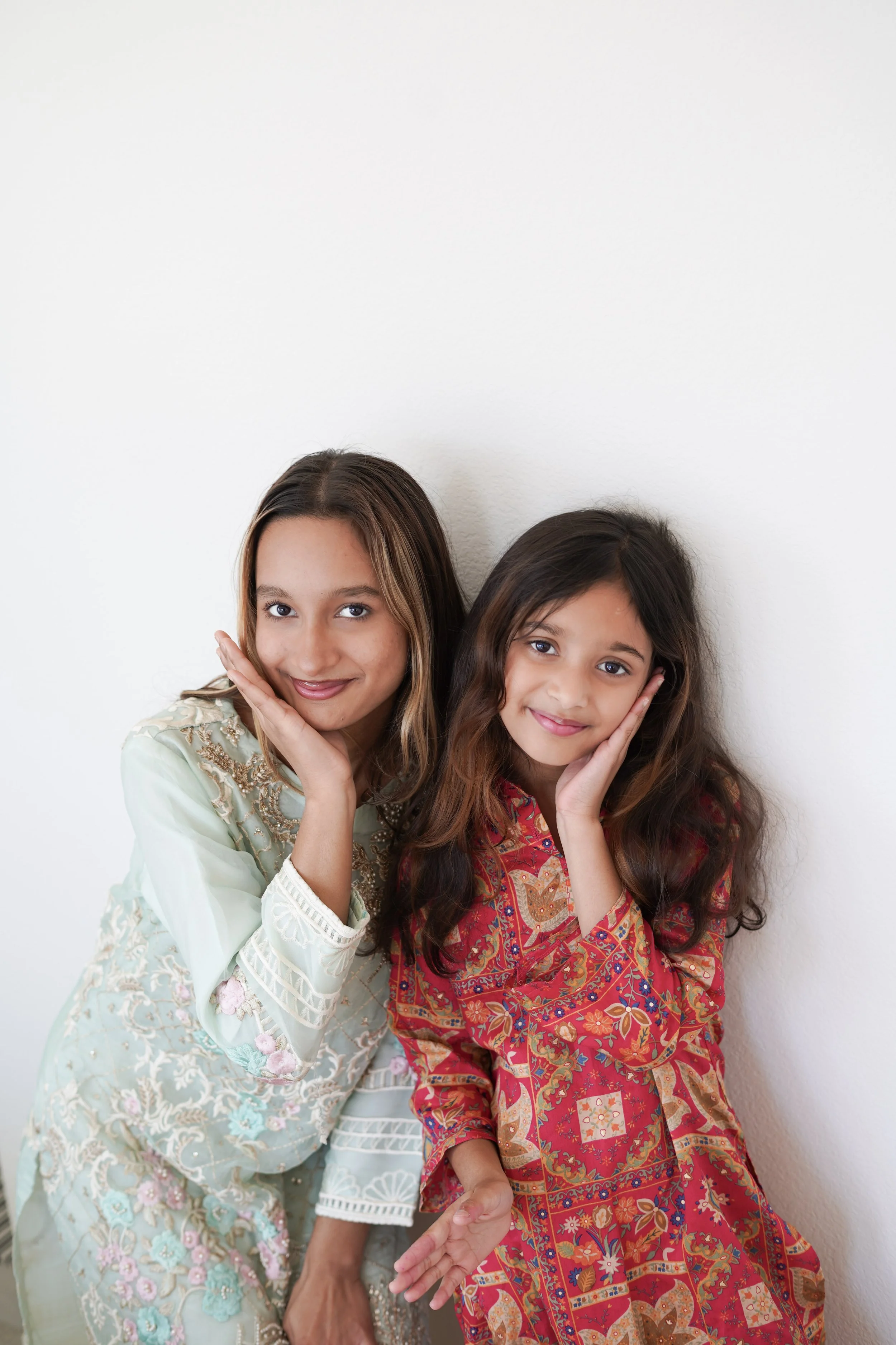 Two young girls wearing colorful, patterned dresses leaning against a white wall, smiling and posing with their hands on their cheeks.