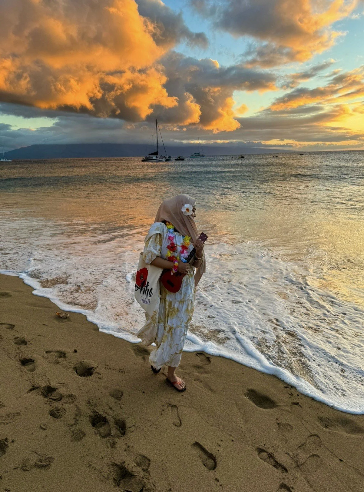 Person wearing a hijab and floral dress walking along the beach at sunset, holding a pink phone and a ukulele, with sailboats in the water and a mountain in the background.