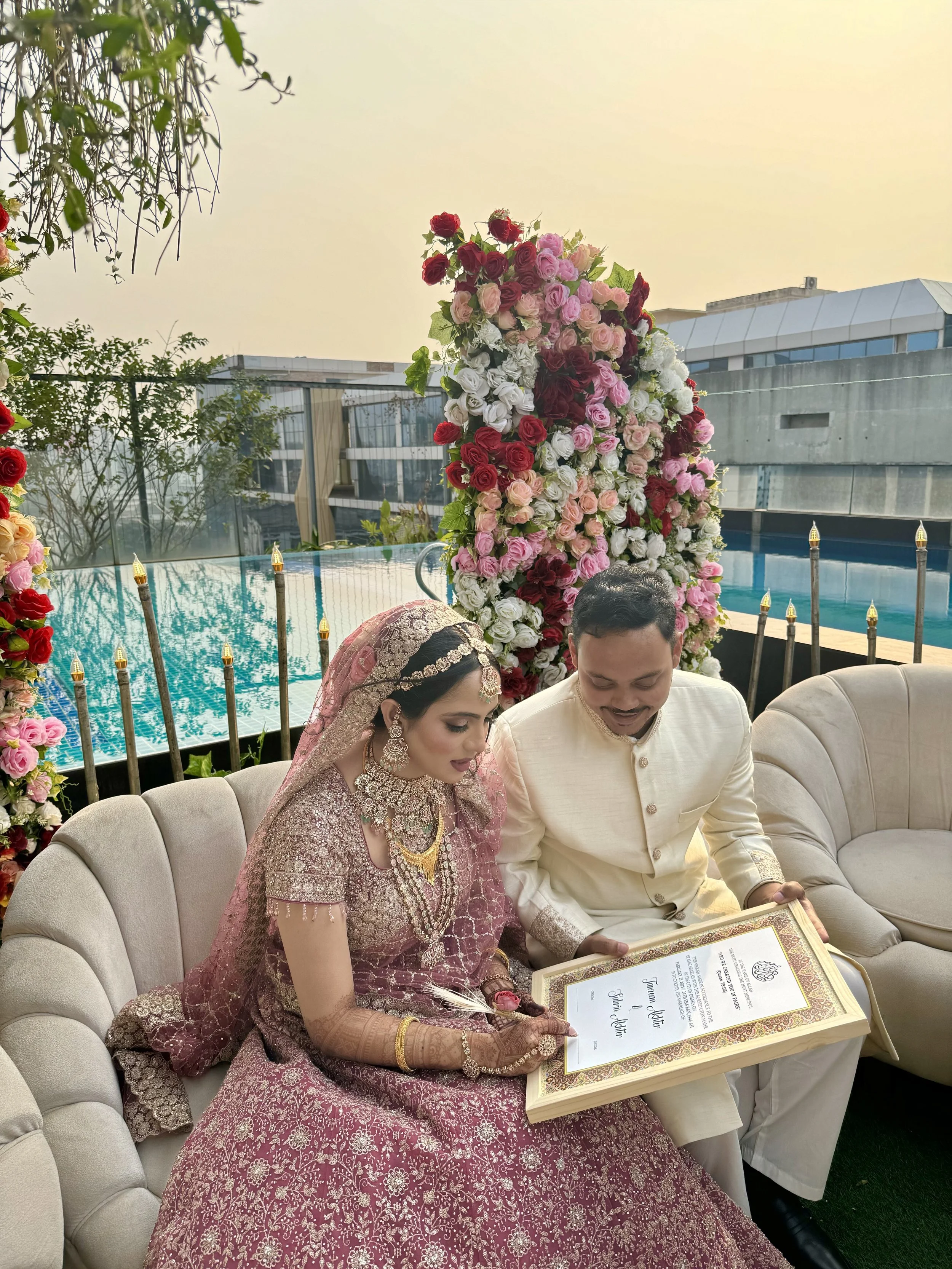 Bride and groom in traditional Indian wedding attire sitting on a cream-colored sofa, signing a marriage document, with a large floral arrangement of roses behind them, near a pool outdoors.