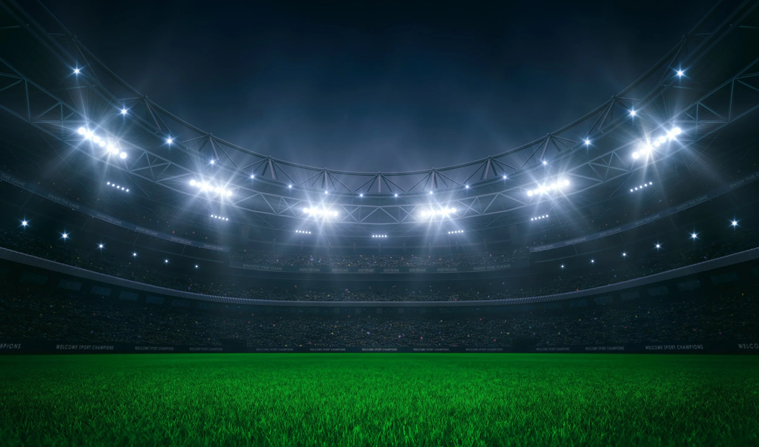 Empty illuminated sports stadium with bright overhead lights and green grass field.