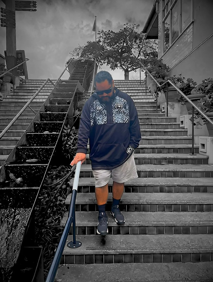 A man with sunglasses, a beard, wearing a black and patterned jacket, white shorts, and sneakers, descending a set of outdoor metal stairs.