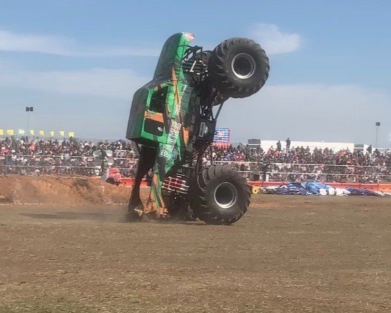 A monster truck upside down with its wheels in the air during a monster truck show on a dirt arena. Spectators in the background watch the event.