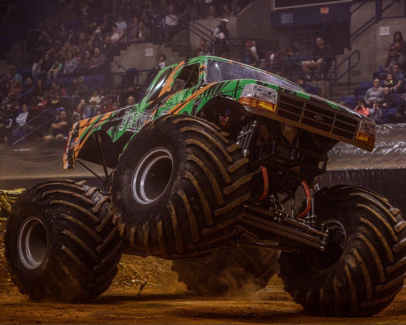Monster truck performing a wheelie during a monster truck event in an indoor arena with spectators watching.