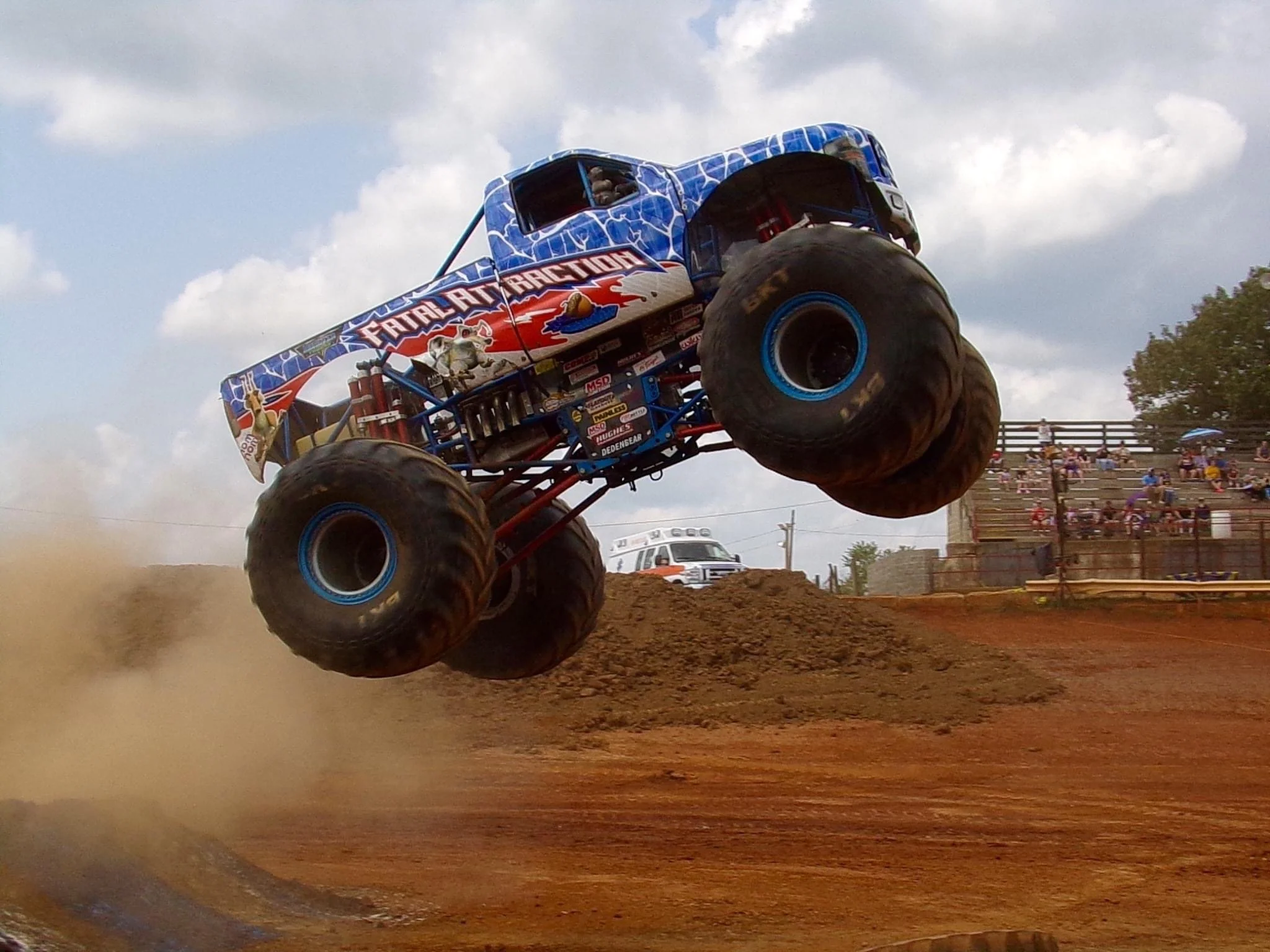 A monster truck with large tires lifting off a dirt track during a race, with spectators watching from bleachers in the background.