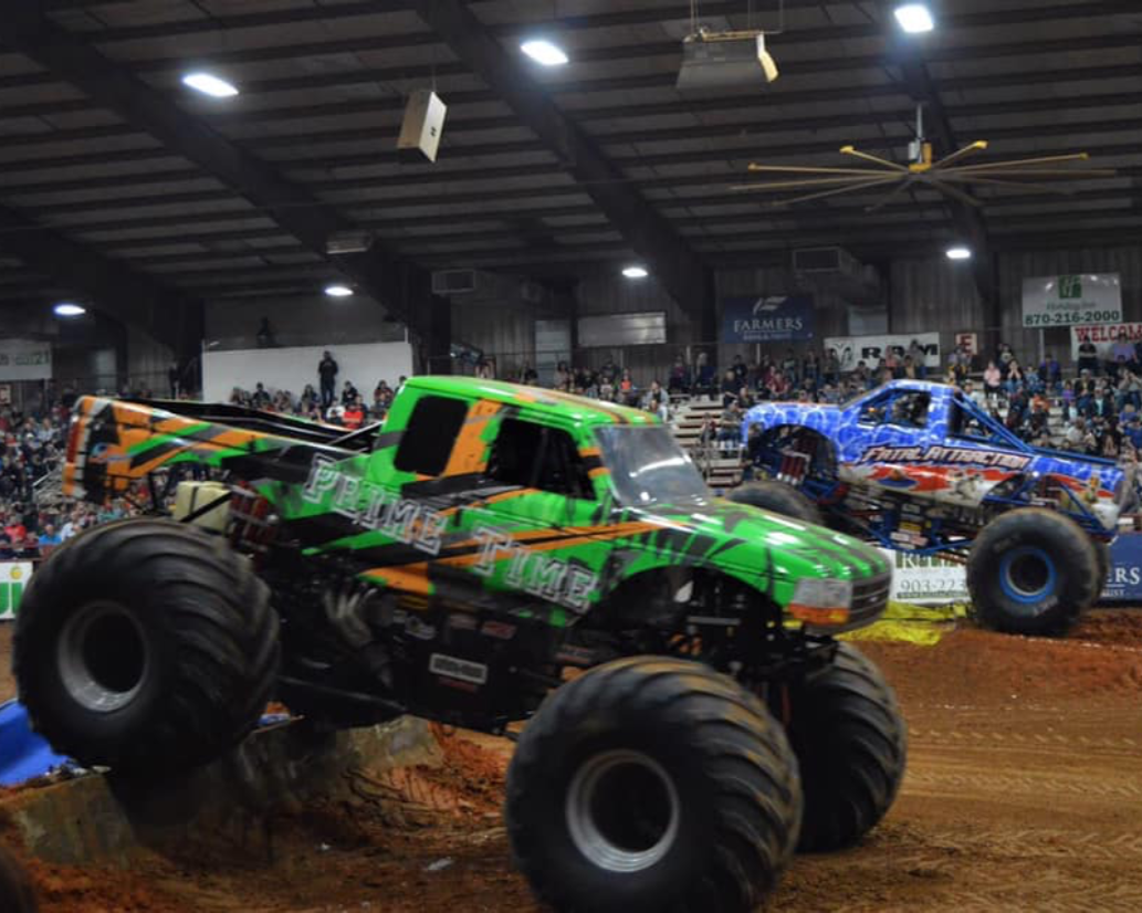 Two monster trucks racing inside an indoor arena with a crowd in the stands and banners hanging on the walls.