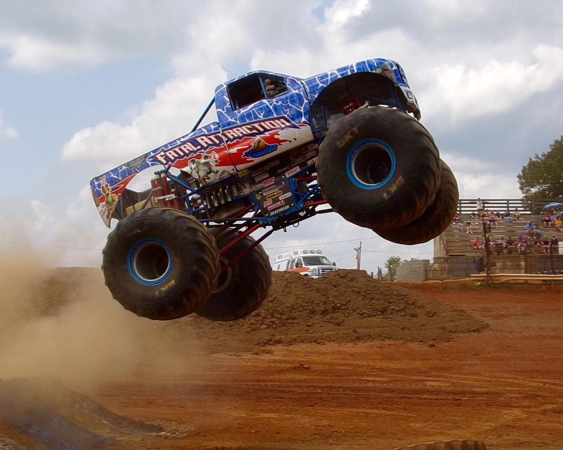 A monster truck with large blue tires and a blue and red paint job jumps over dirt at an outdoor event. Bleachers with spectators are visible in the background under a partly cloudy sky.