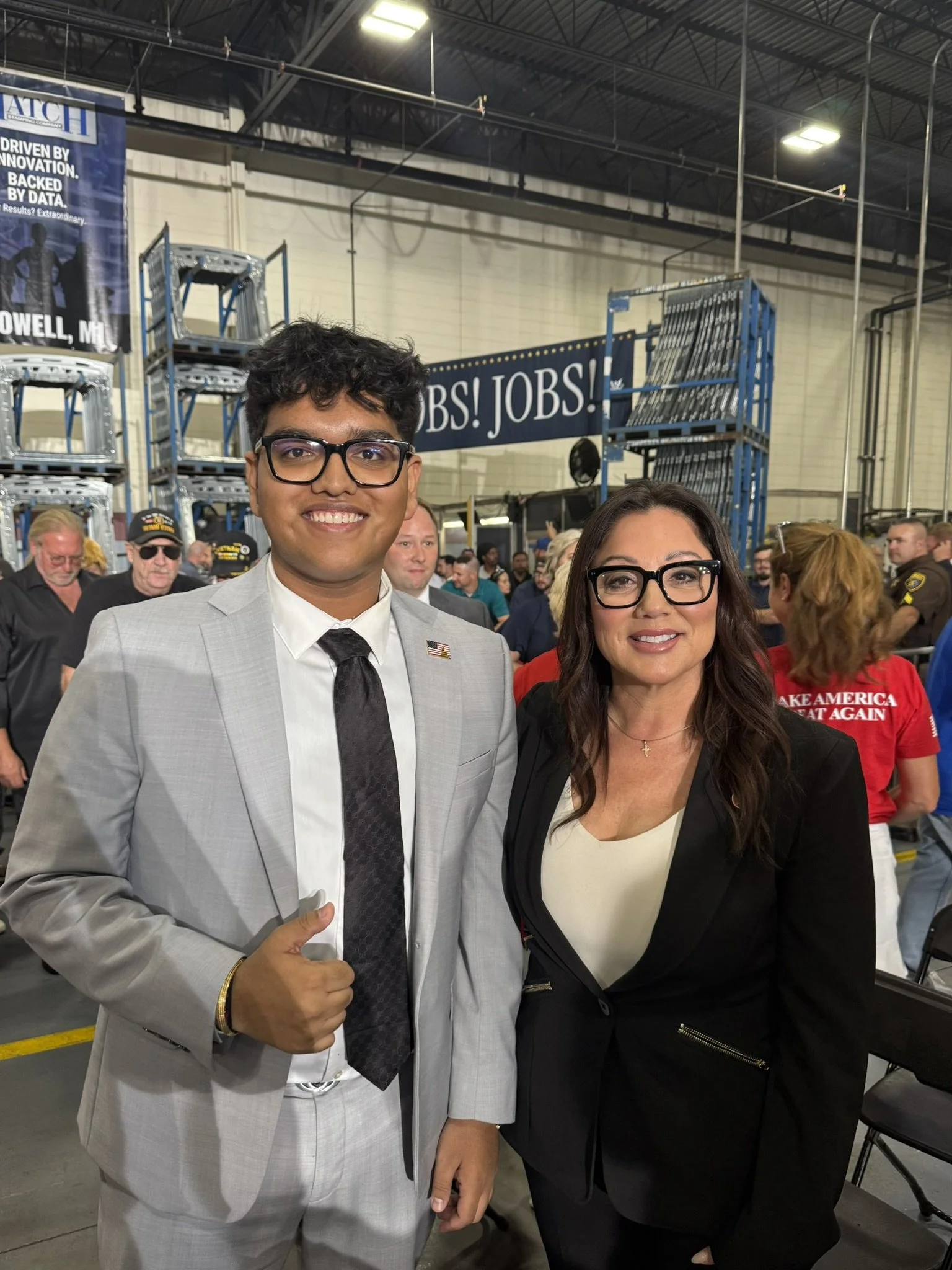 Two people standing together at a political event, surrounded by a crowd. The man on the left is wearing a light gray suit, white shirt, dark patterned tie, glasses, and giving a thumbs up. The woman on the right is wearing a black blazer, white top,