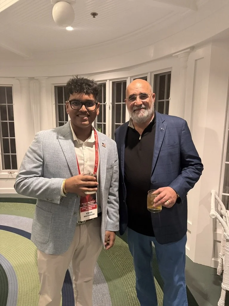 Two men standing together indoors at a social event, holding drinks, with a white ceiling and large windows in the background.