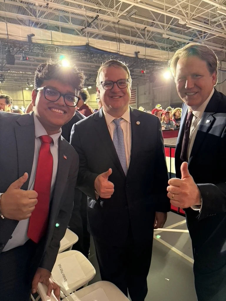 Three men dressed in suits smiling and giving thumbs up at an indoor event, with a crowd and bright lighting in the background.